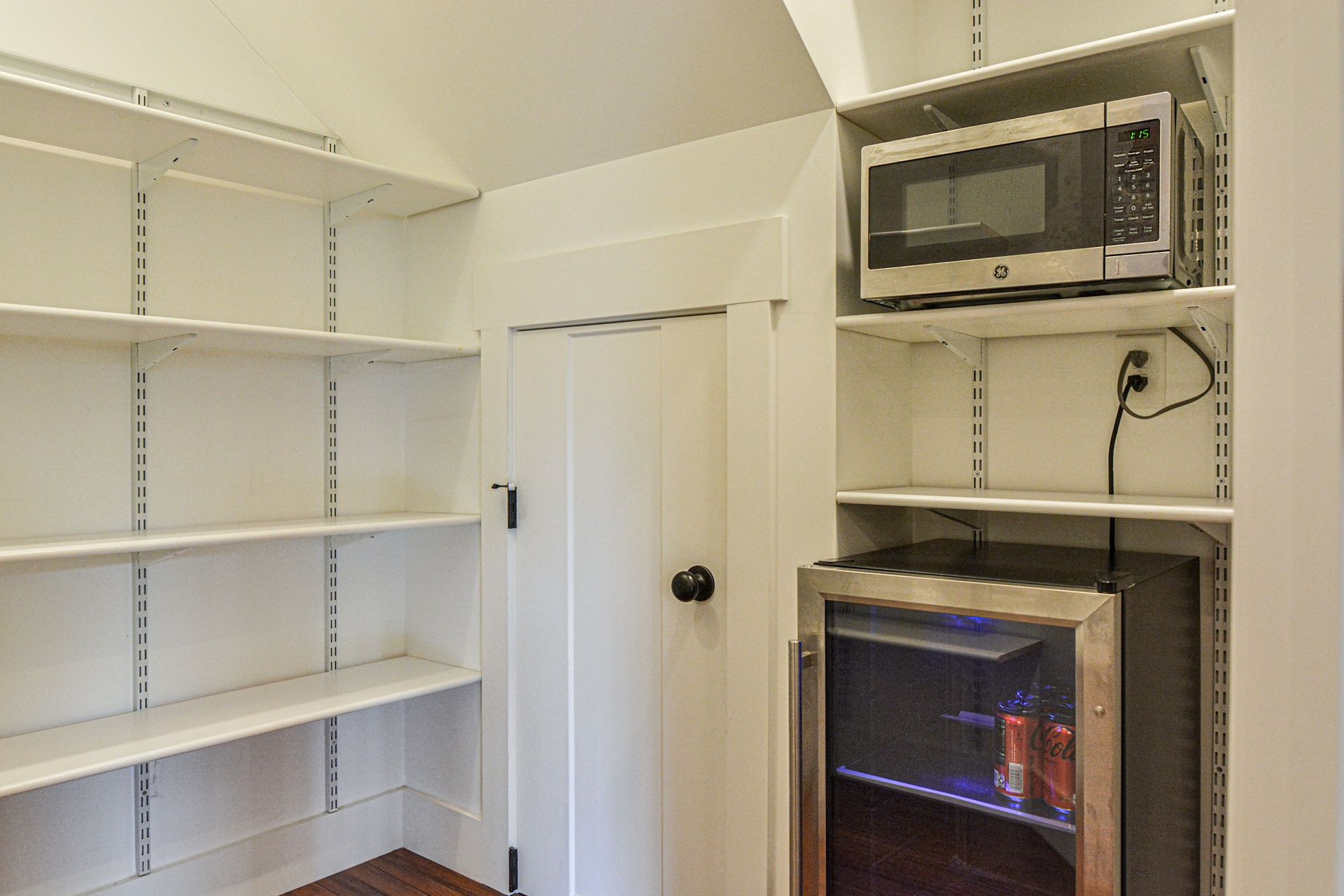White pantry shelves, a door, a microwave on a shelf, and a small glass-front beverage refrigerator.