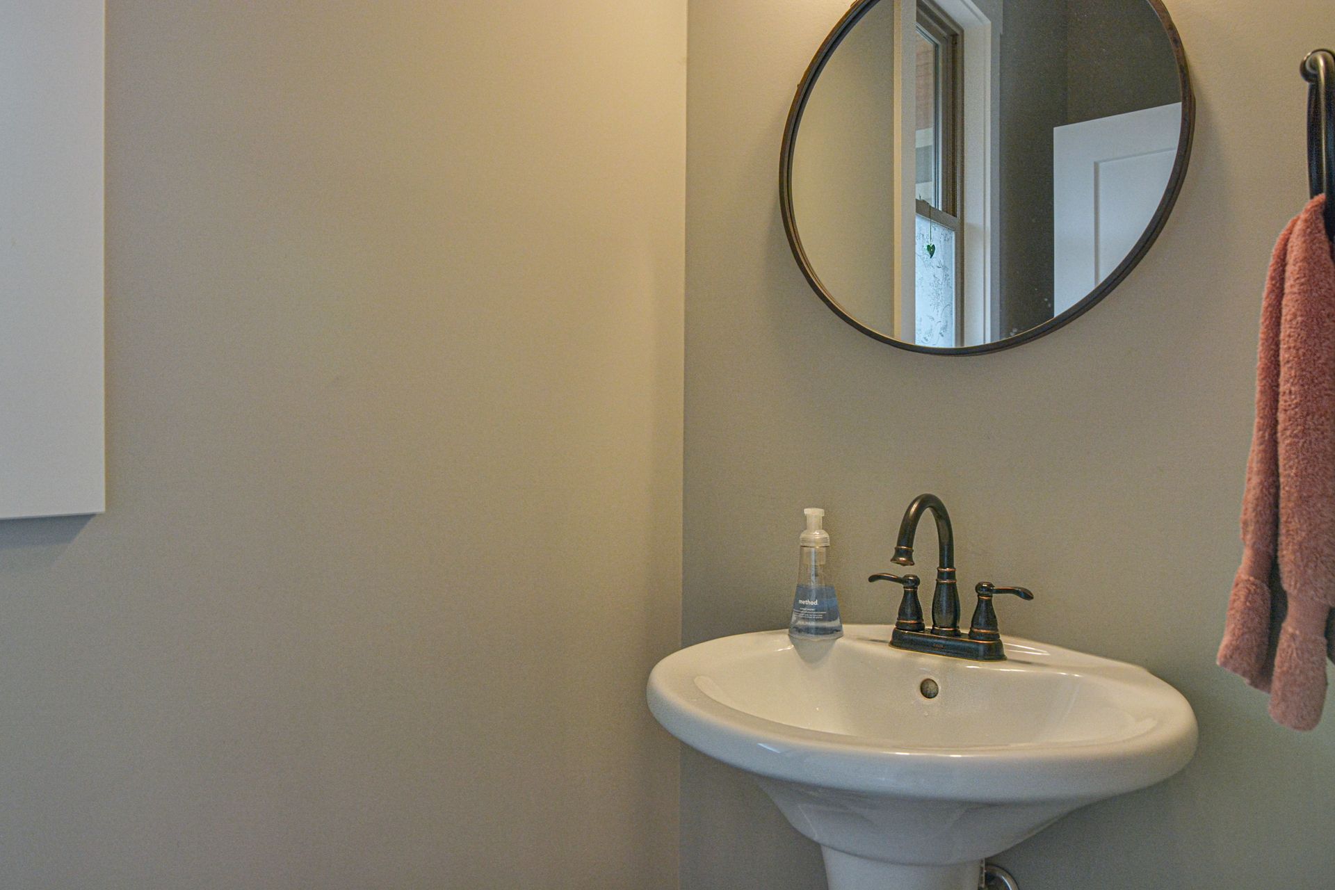 A pedestal sink with a dark bronze faucet and soap dispenser beneath a circular mirror on a light grey bathroom wall.