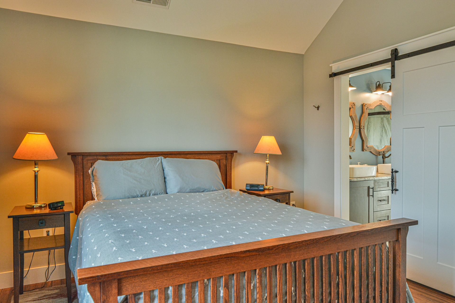 A bedroom with a wooden bed frame, blue bedding, bedside lamps, and a white sliding barn door opening to a bathroom.