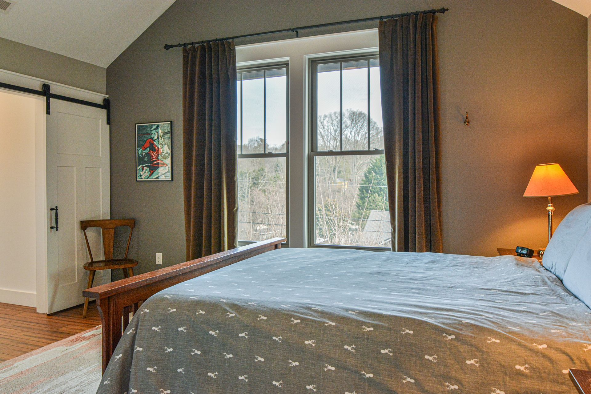A cozy bedroom with a bed, a wooden chair in the corner, dark curtains over two windows, and a warm bedside lamp.