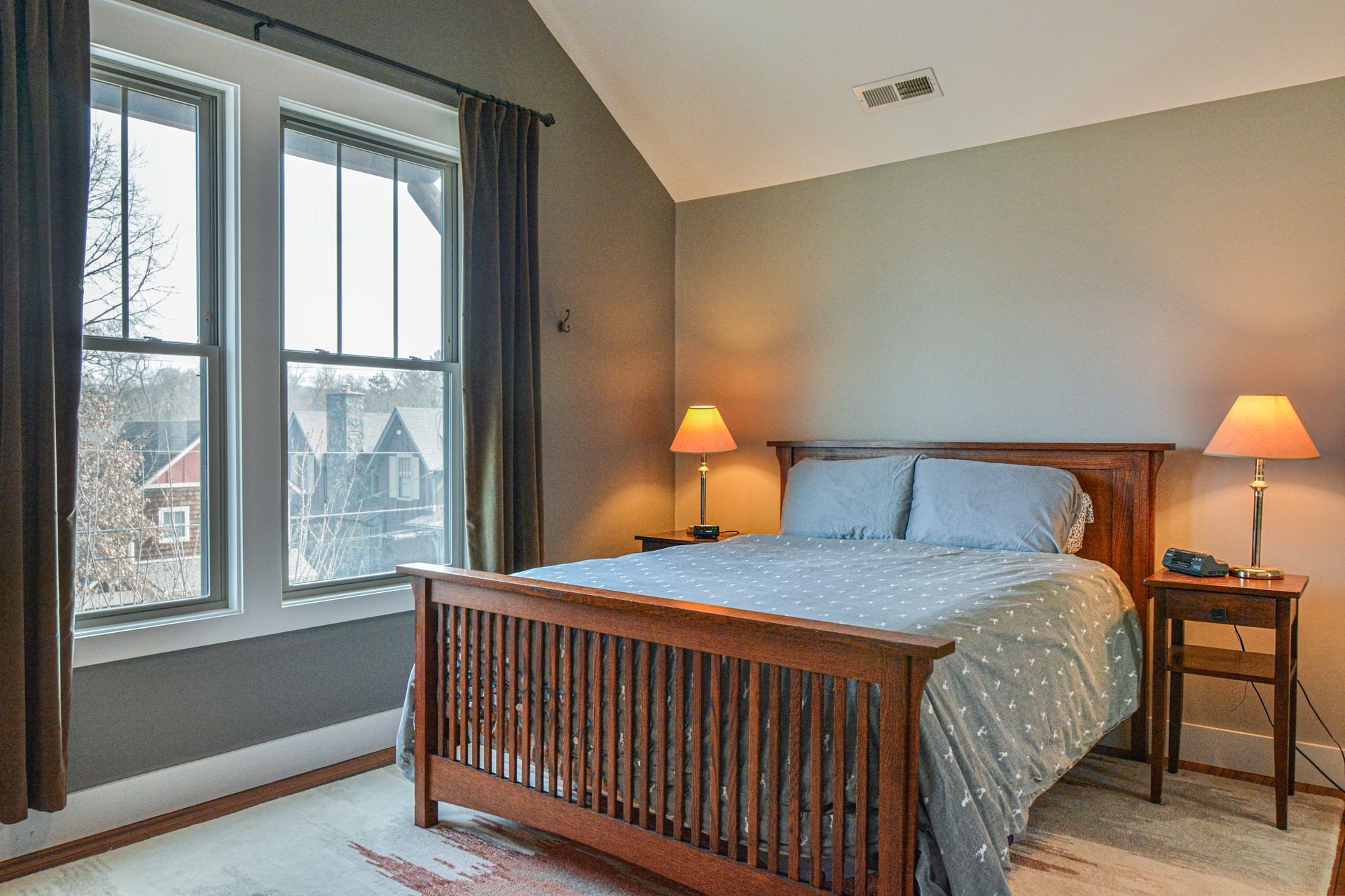 A bedroom with a wooden bed frame, blue polka-dot bedding, two lamps on nightstands, and two windows with brown curtains.