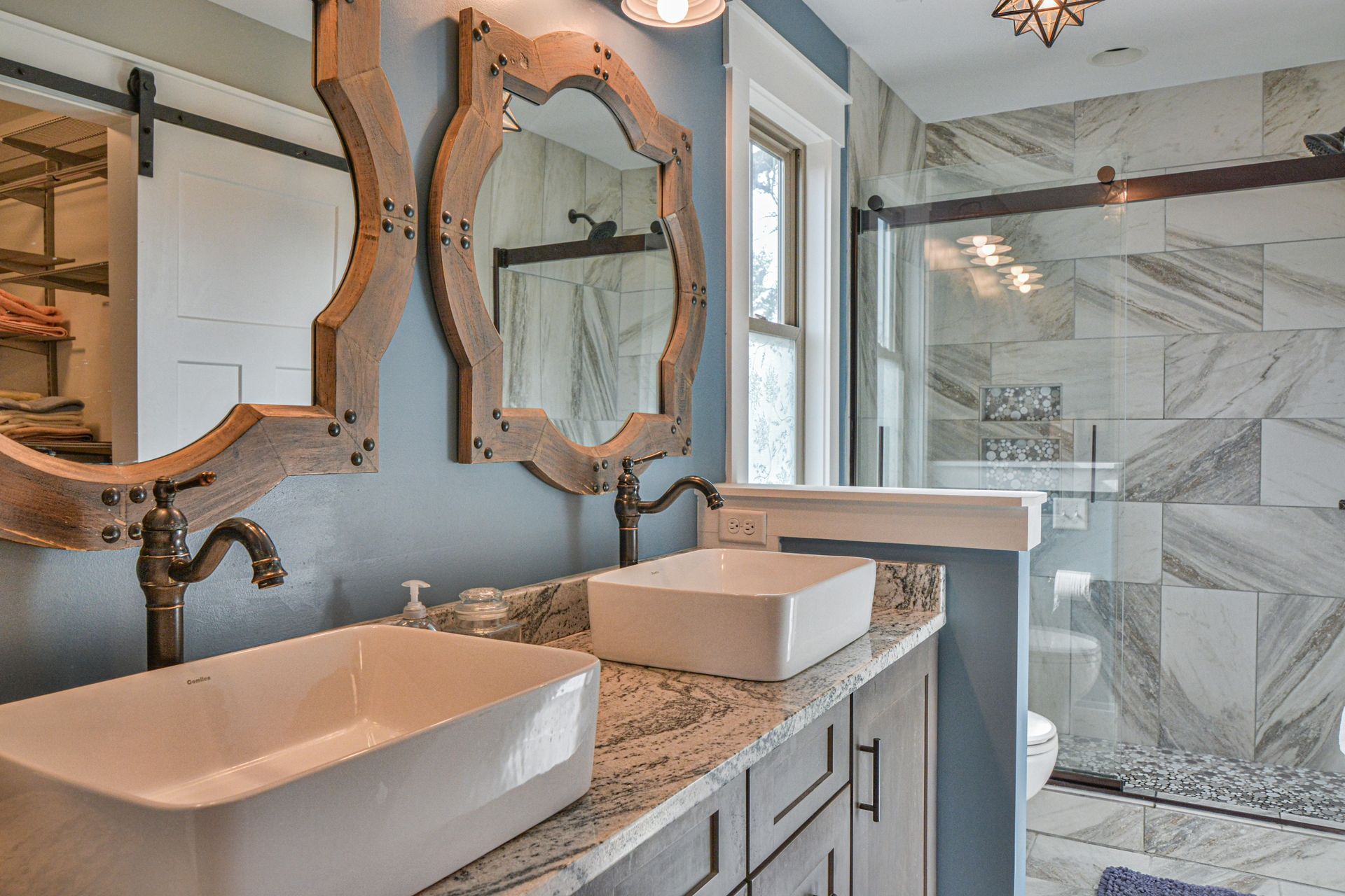 Double vessel sinks on a granite vanity with ornate wooden mirrors in a bathroom with a marble walk-in shower.