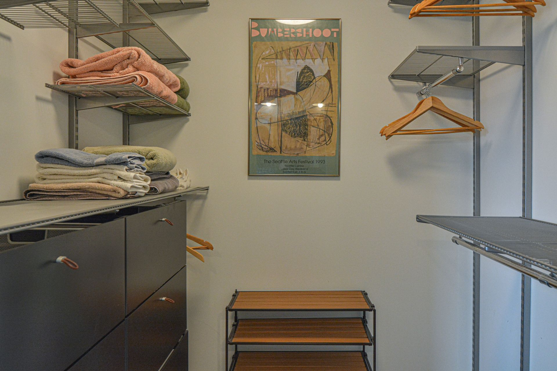 A walk-in closet featuring metal shelving units with folded towels, hanging clothes, and a small wooden shoe rack.