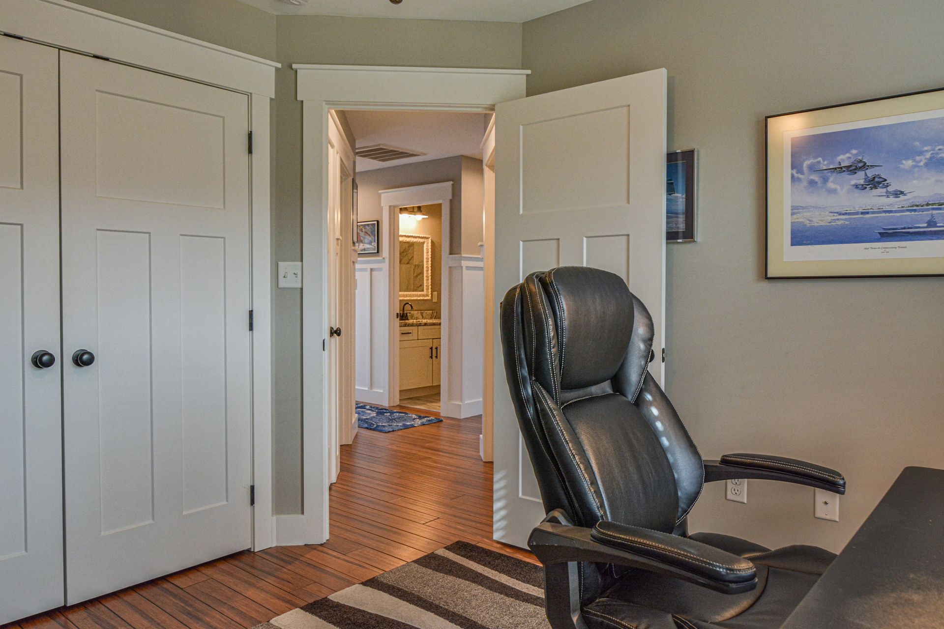 An office space with a black leather chair facing a desk, beside double doors and a view into a bathroom down the hall.