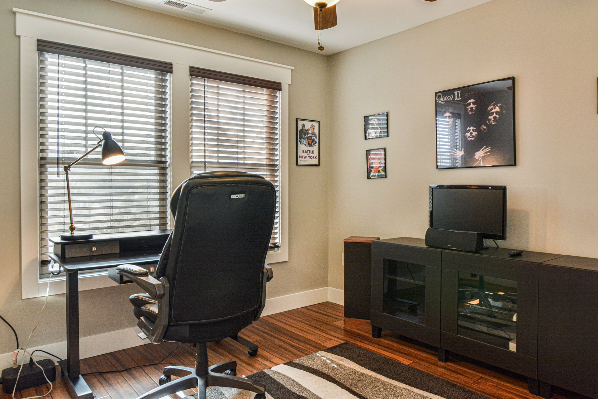 A home office featuring a desk with a lamp, a black office chair, a media console with a TV, and framed wall art.