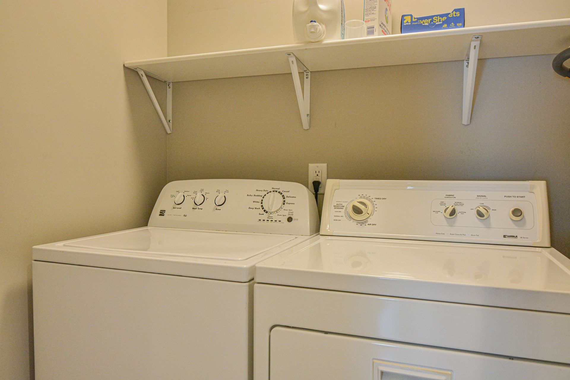 A white washing machine and clothes dryer side-by-side against a beige wall, beneath a mounted white storage shelf.