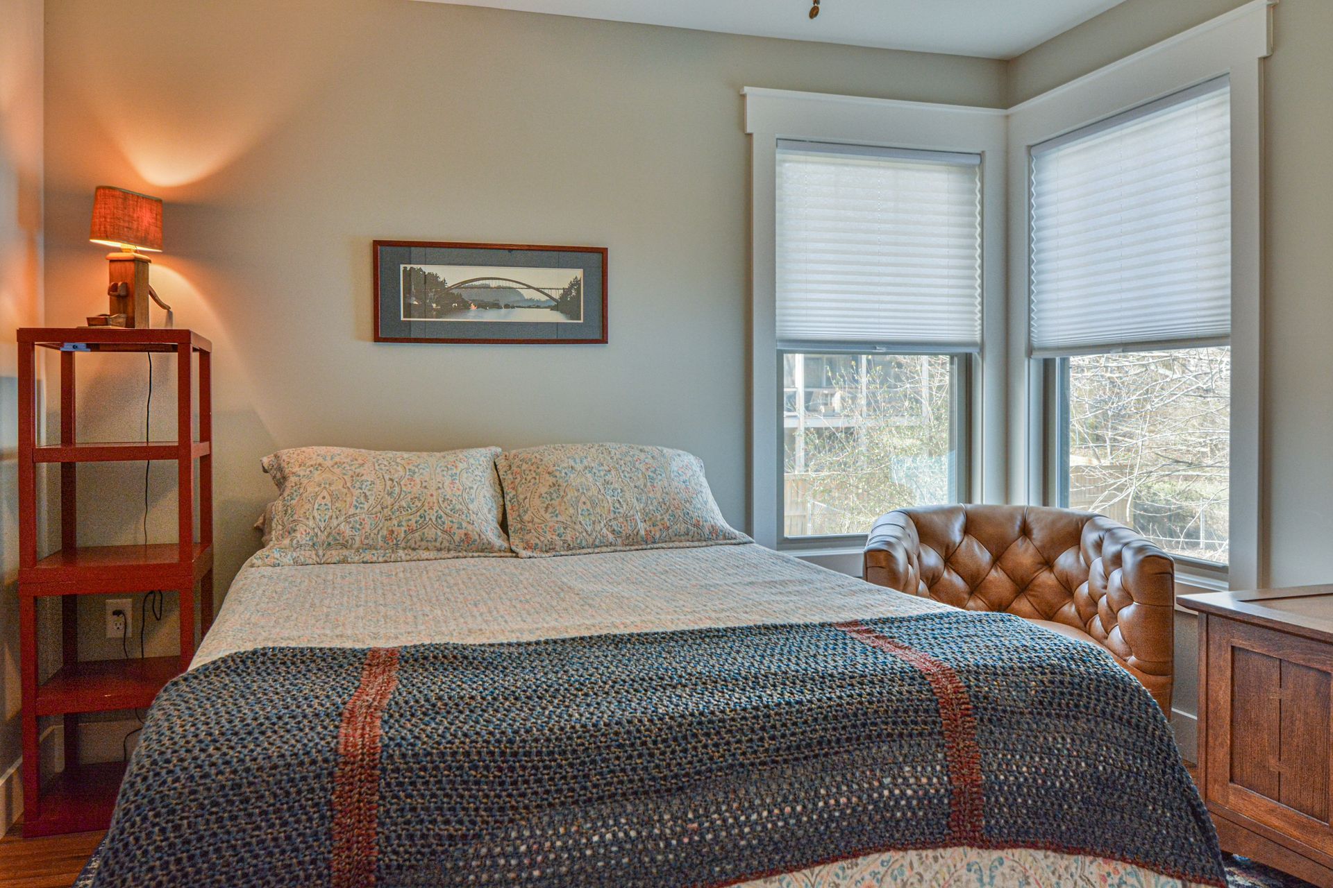 A bedroom featuring a bed with a textured quilt, a tufted leather chair, a red shelving unit, and windows with blinds.