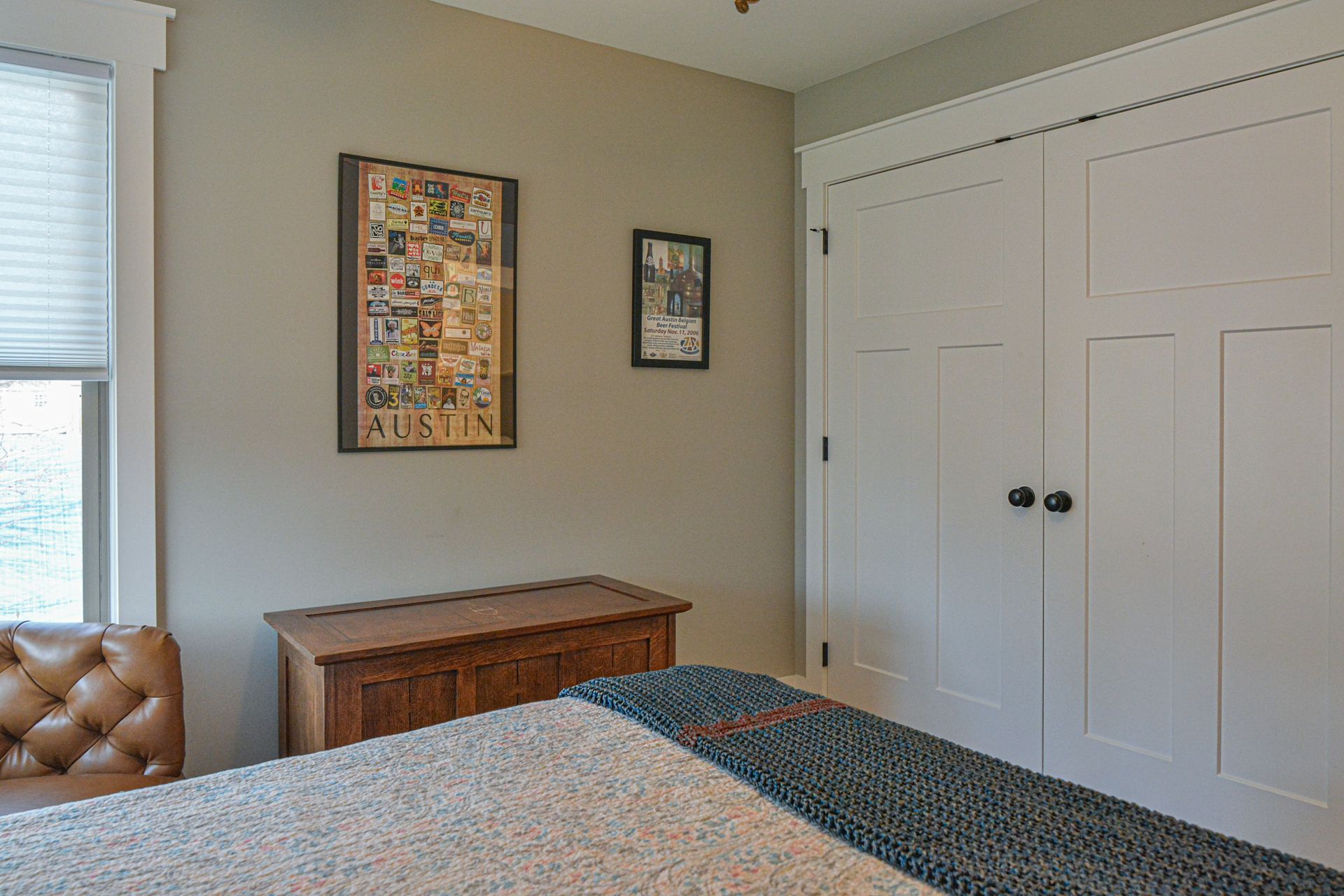 A corner of a bedroom with a tan wall, two framed pictures, a wooden trunk, a tufted chair, and white double doors.
