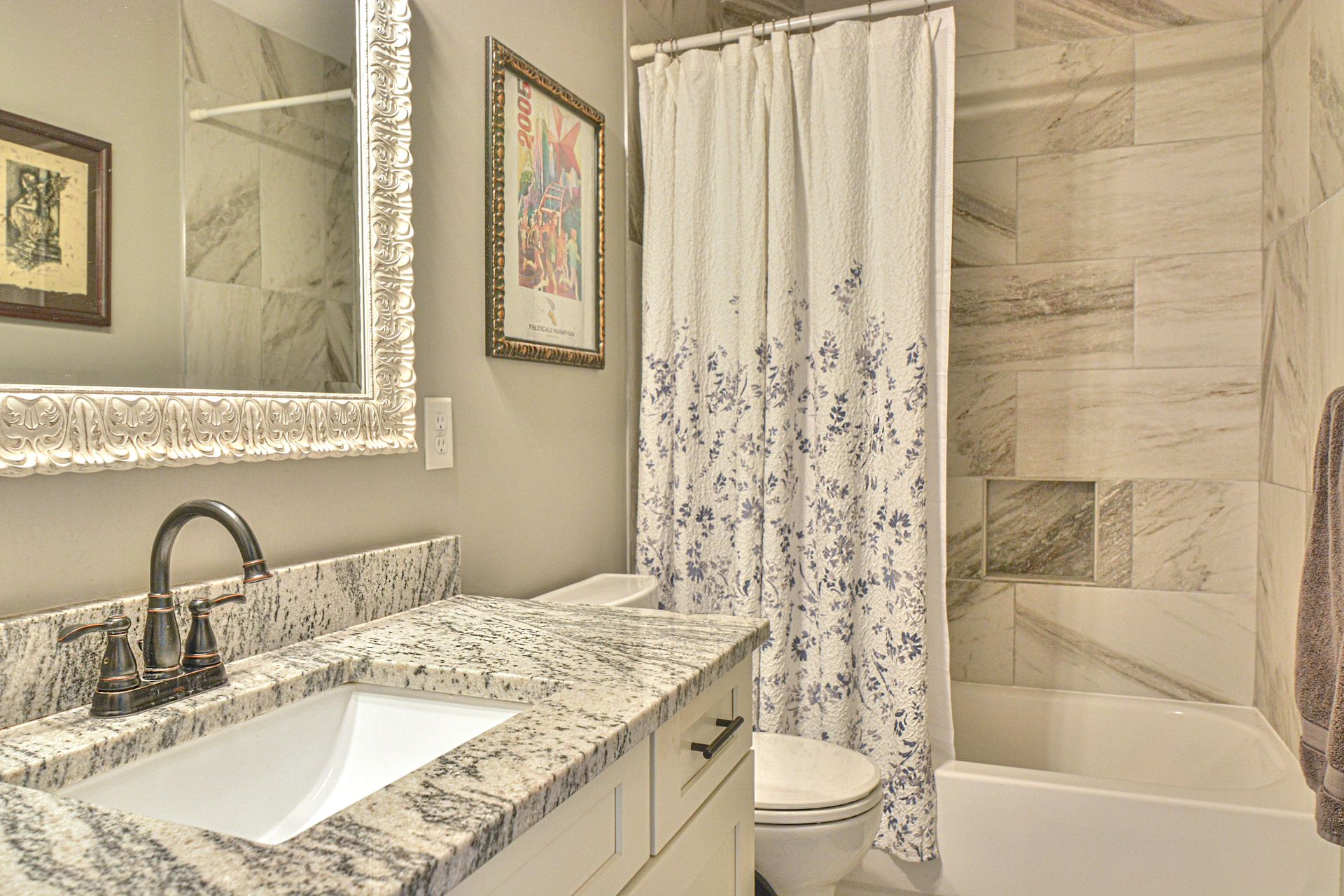 A bathroom with granite countertops, a framed mirror, a patterned shower curtain, and a tiled shower-tub combo.