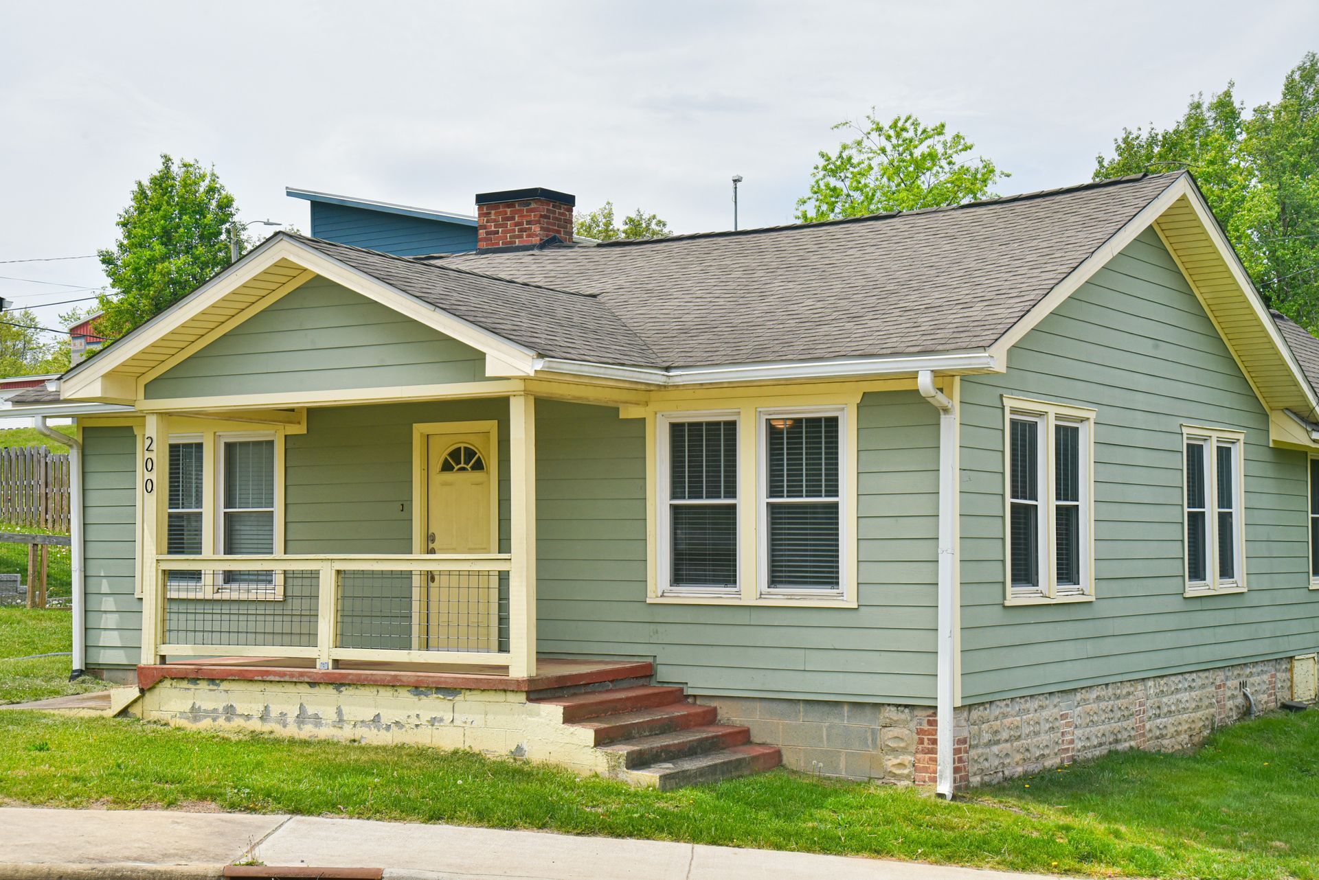 Small green bungalow house with a covered porch, white trim, and brick steps in a suburban yard