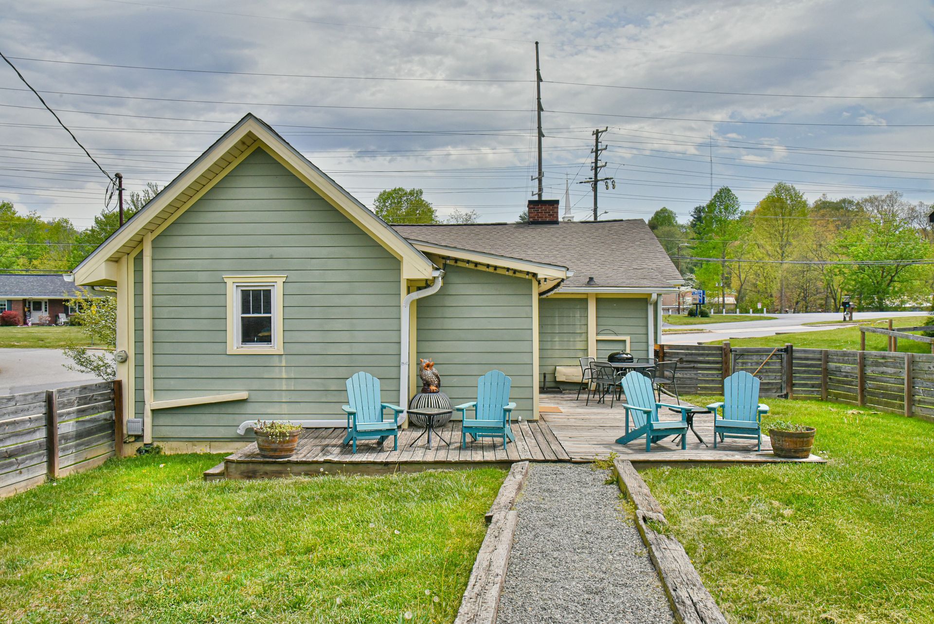 Small green house with a front lawn, stone path, and blue chairs on a patio under a cloudy sky
