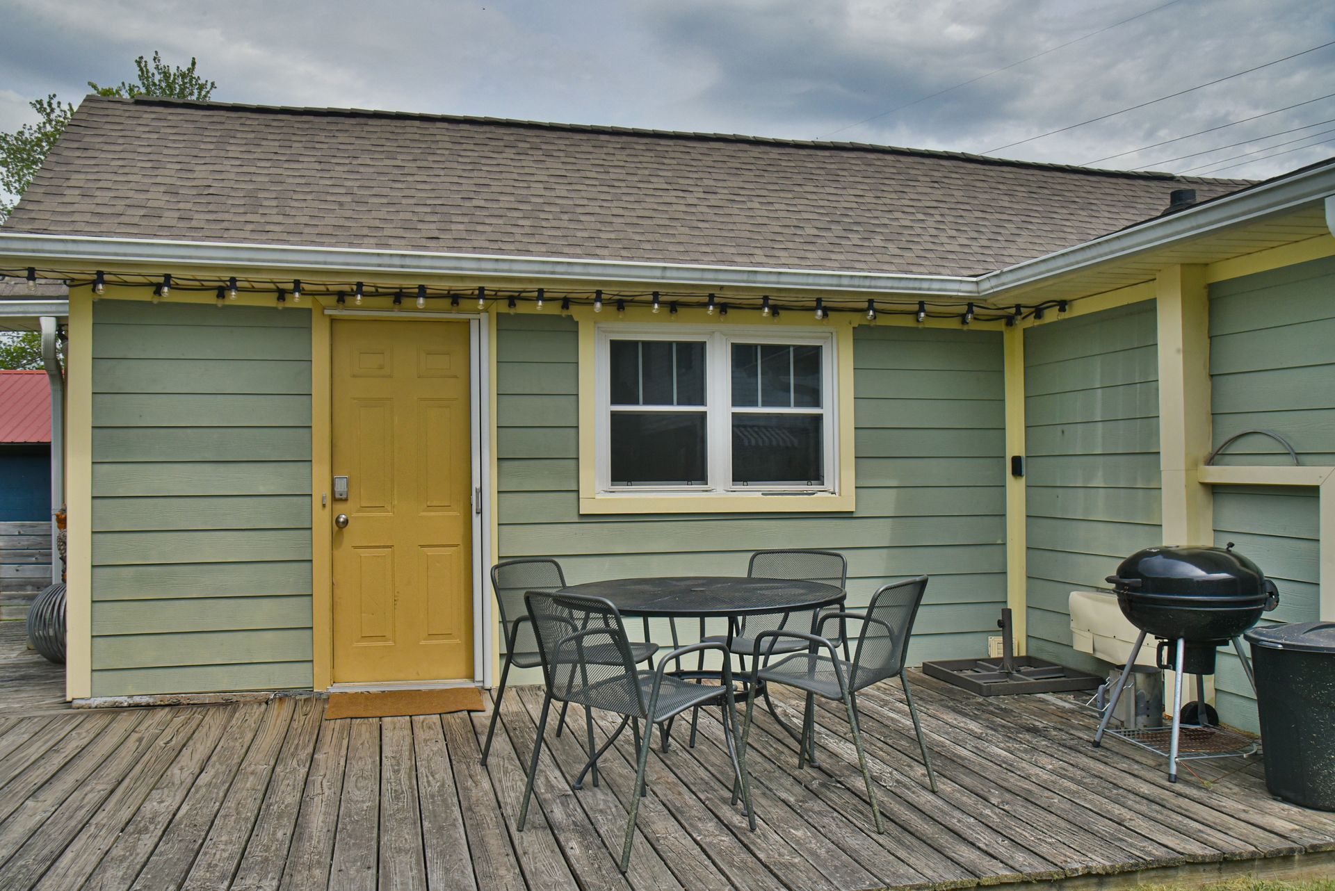 Small wooden porch with green siding, yellow door, window, patio table, chairs, and a grill.