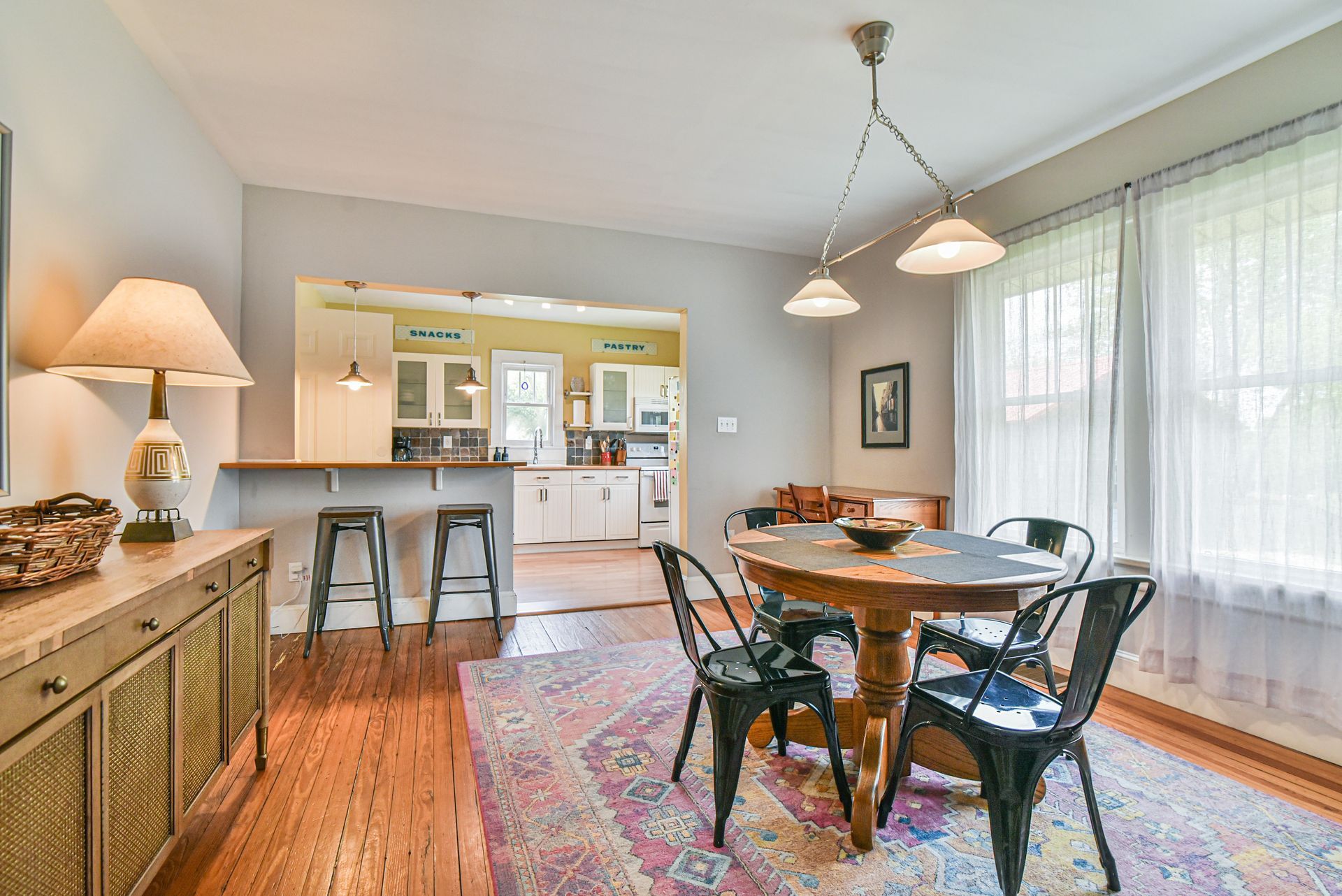 Bright dining room with round table, chairs, patterned rug, and open view to a kitchen counter with stools.