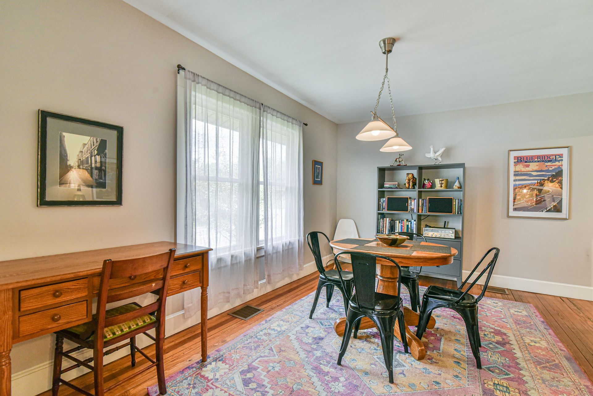 Dining room with wooden table and chairs, colorful rug, bench, and large window with sheer curtains.