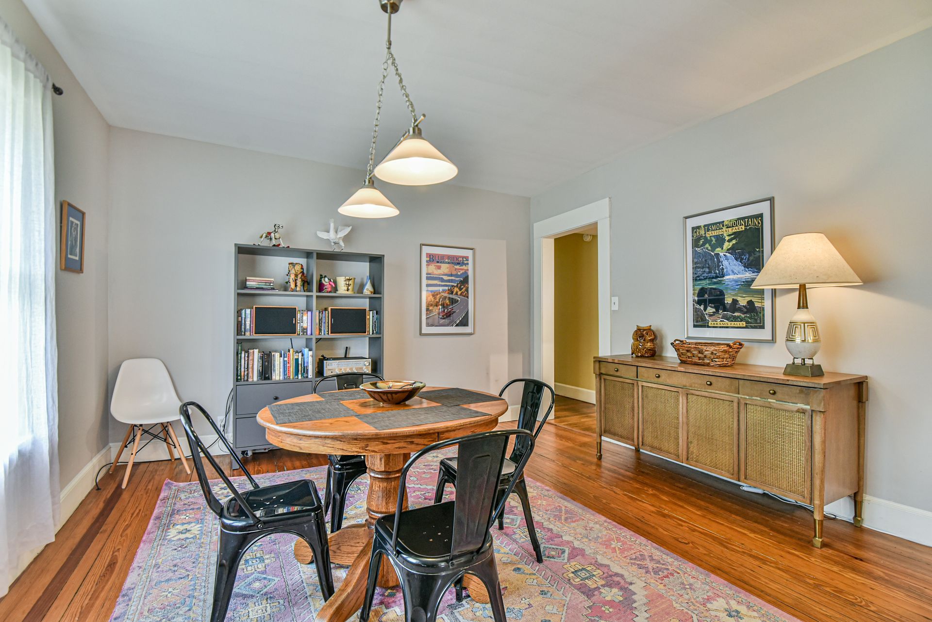 Dining room with round wooden table, black chairs, colorful rug, and sideboard under framed art.