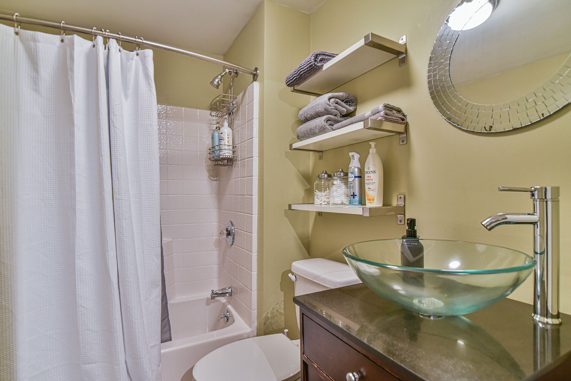 Small bathroom with bathtub, shower curtain, glass sink, shelves, and mirror