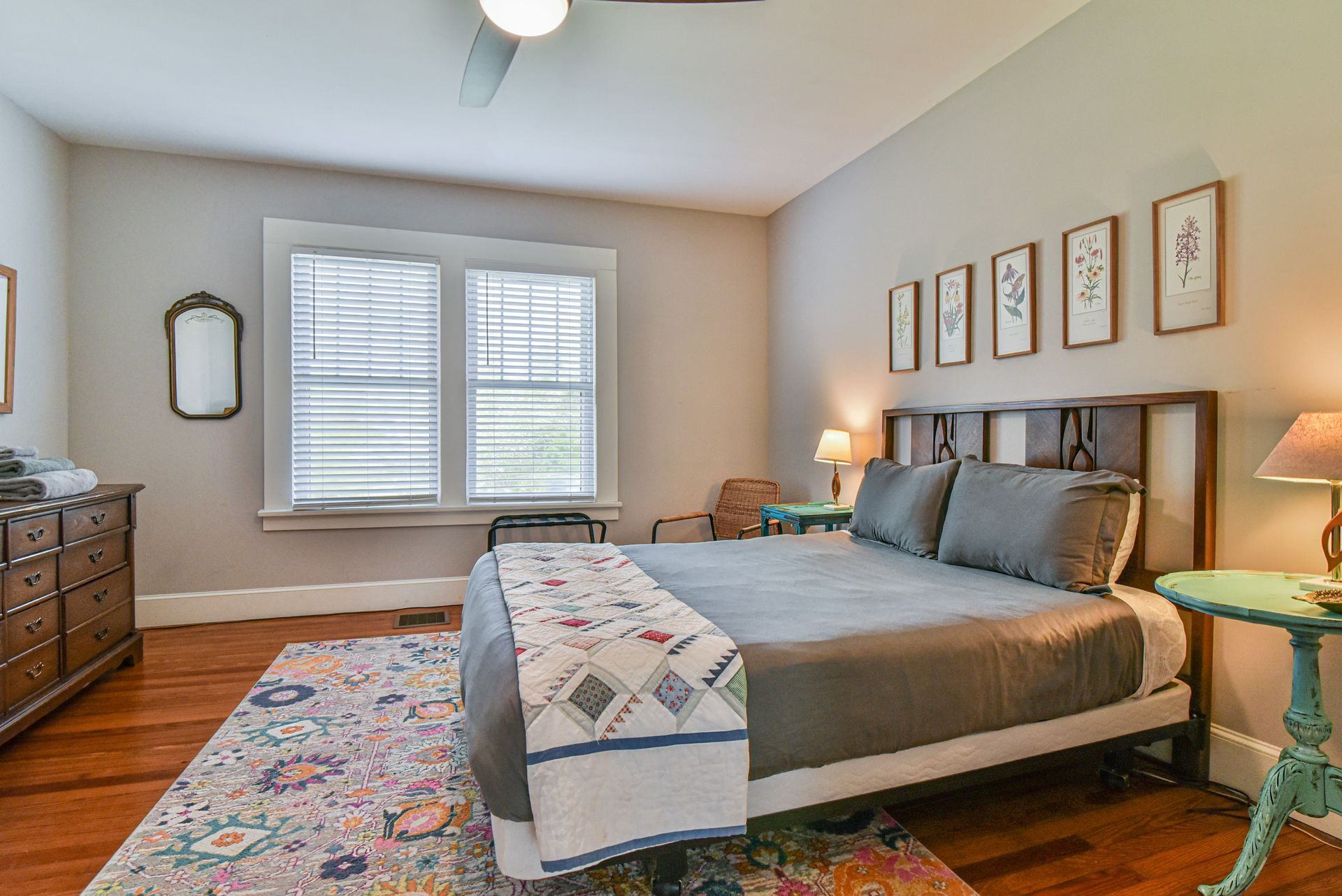 Bright bedroom with a gray bed, patterned rug, white shutters, and framed wall art