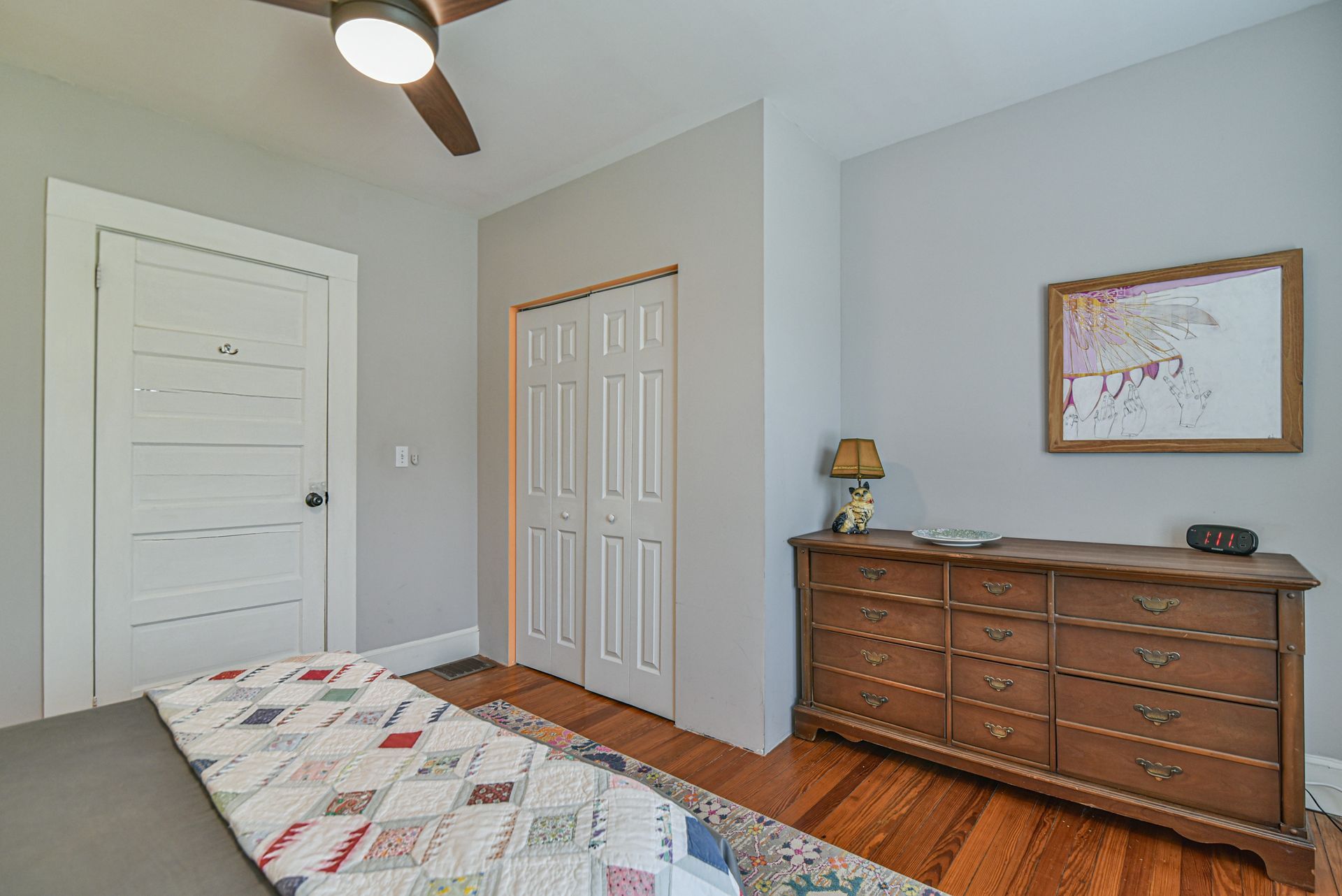 Small bedroom with gray walls, wood floor, dresser, closet doors, and a bed corner in the foreground