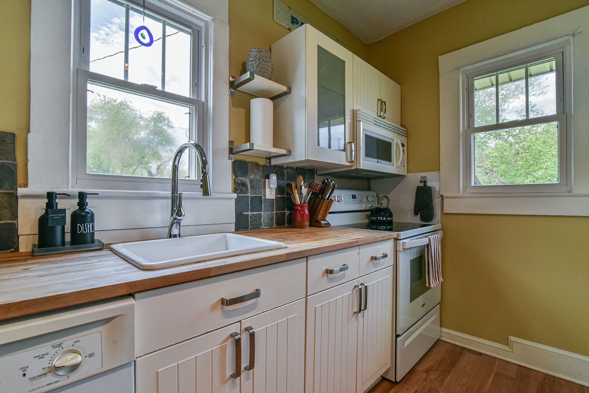 Bright kitchen with white cabinets, wooden countertops, a sink by two windows, and a microwave.