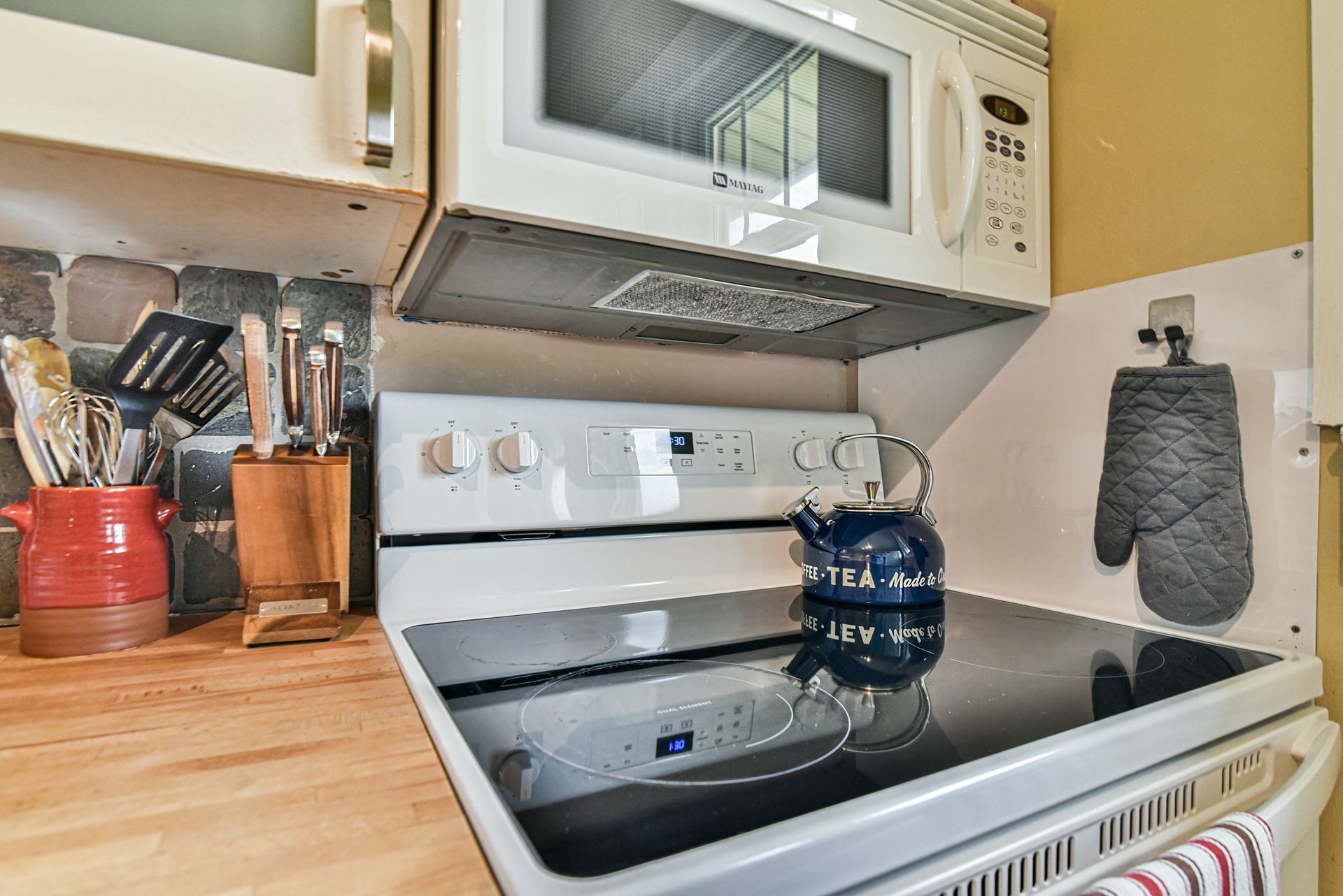 Compact kitchen stove with microwave, wooden countertop, utensils, and a blue kettle on the burner