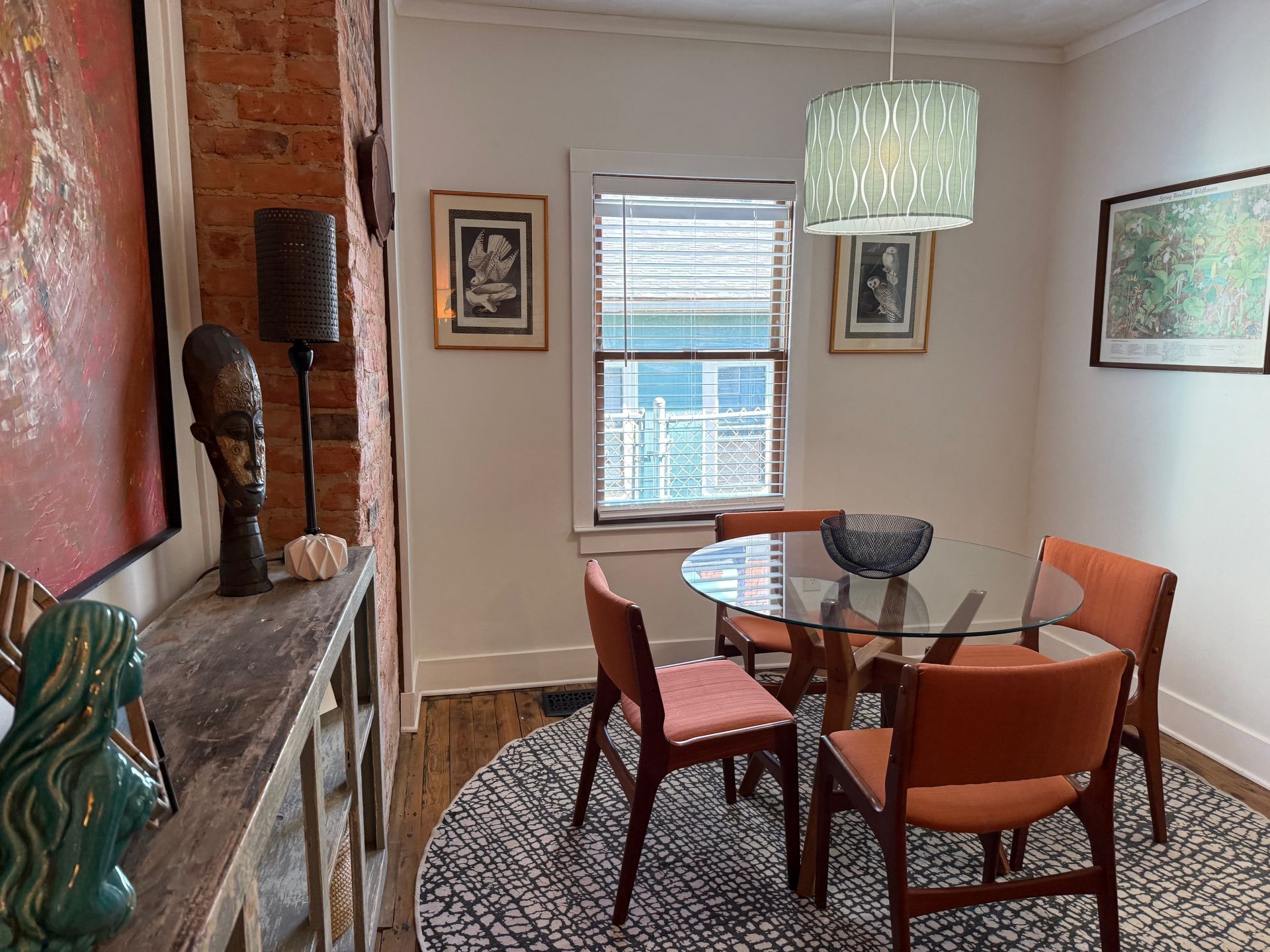 Small dining room with round glass table, orange chairs, window, fireplace, and wall art