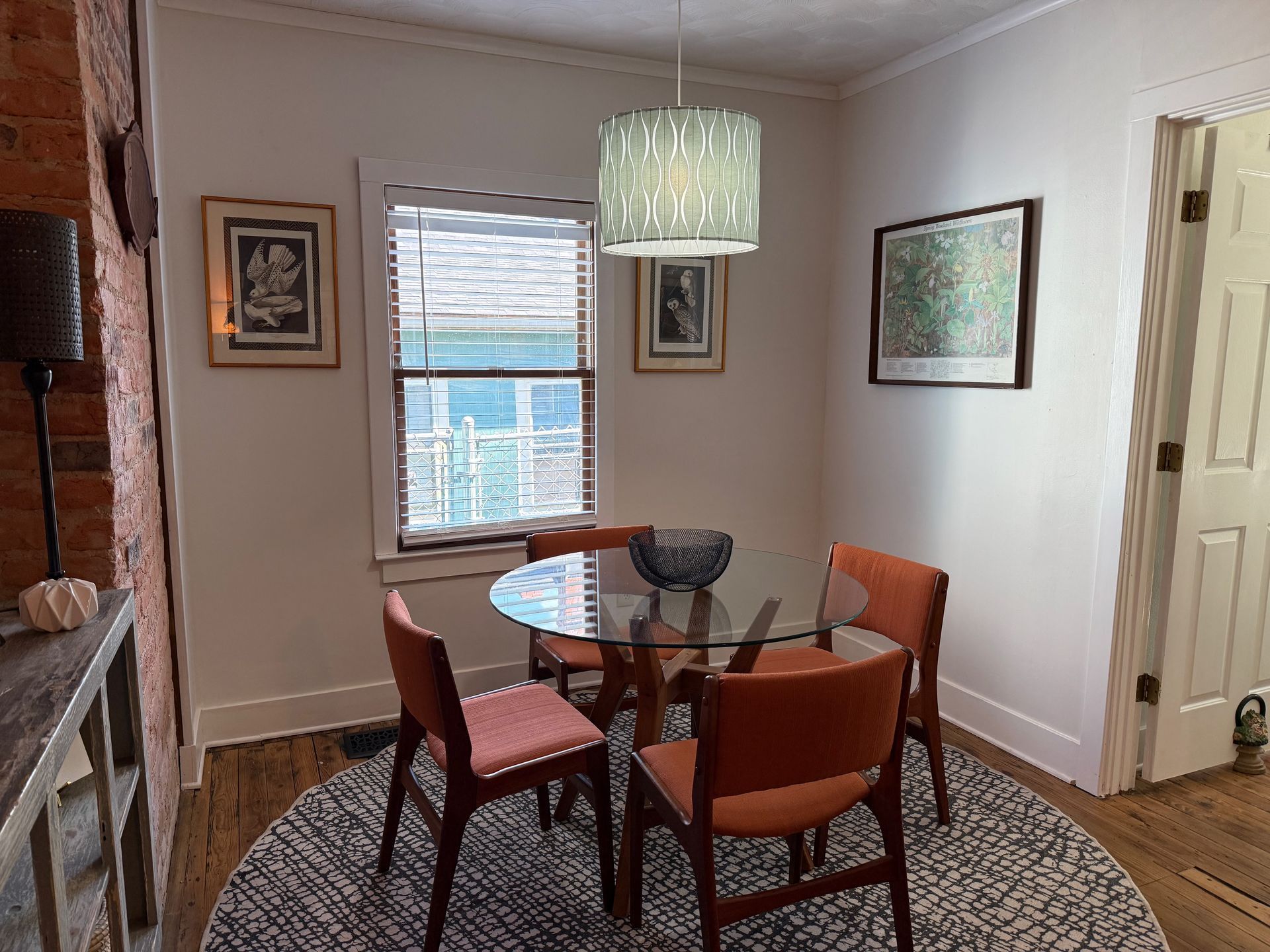 Dining nook with round glass table, four orange chairs, and a green pendant light by a window