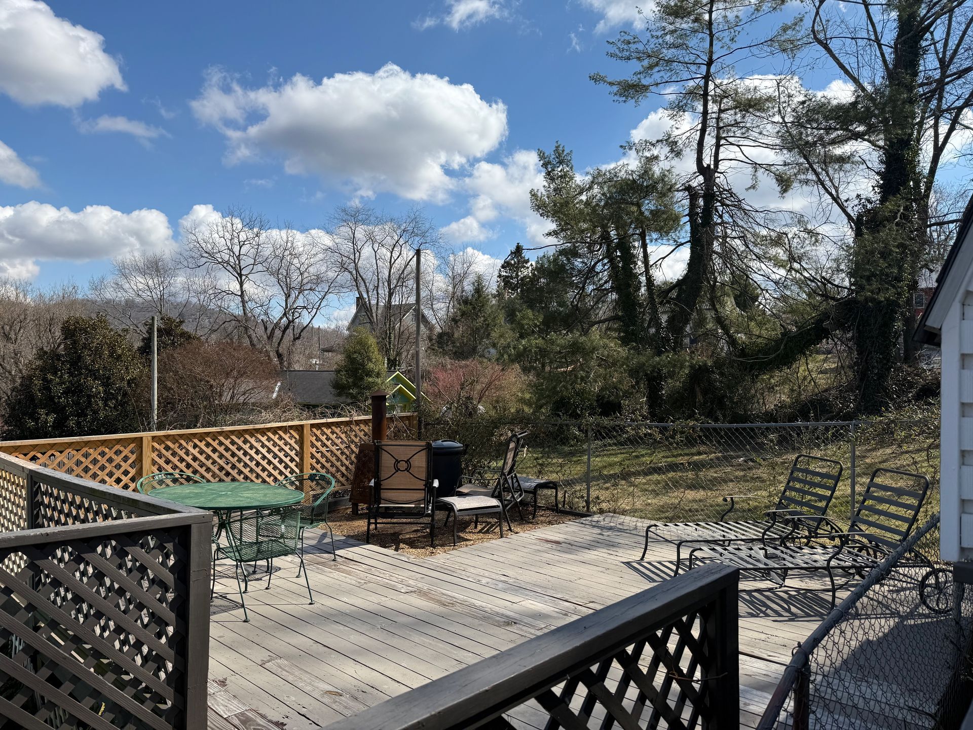 Sunny backyard deck with lattice railing, chairs, and trees under a blue sky with clouds
