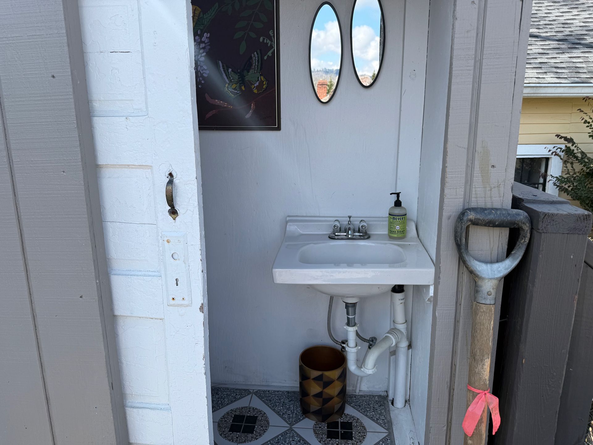 Small outdoor sink beside a doorway, with a soap dispenser and cleaning items on the floor.