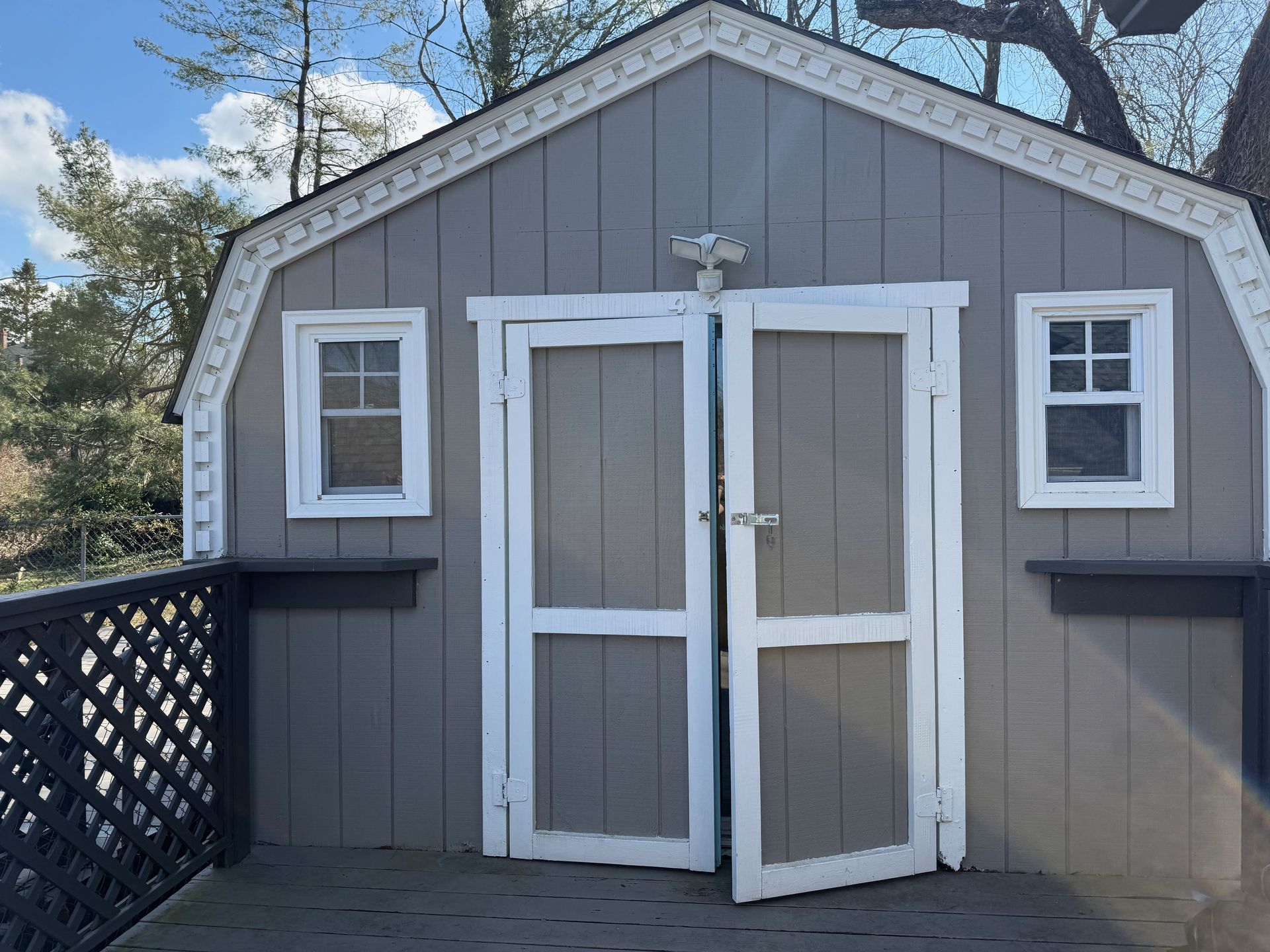 Gray backyard shed with white trim and double doors, one door slightly open, beside a lattice fence