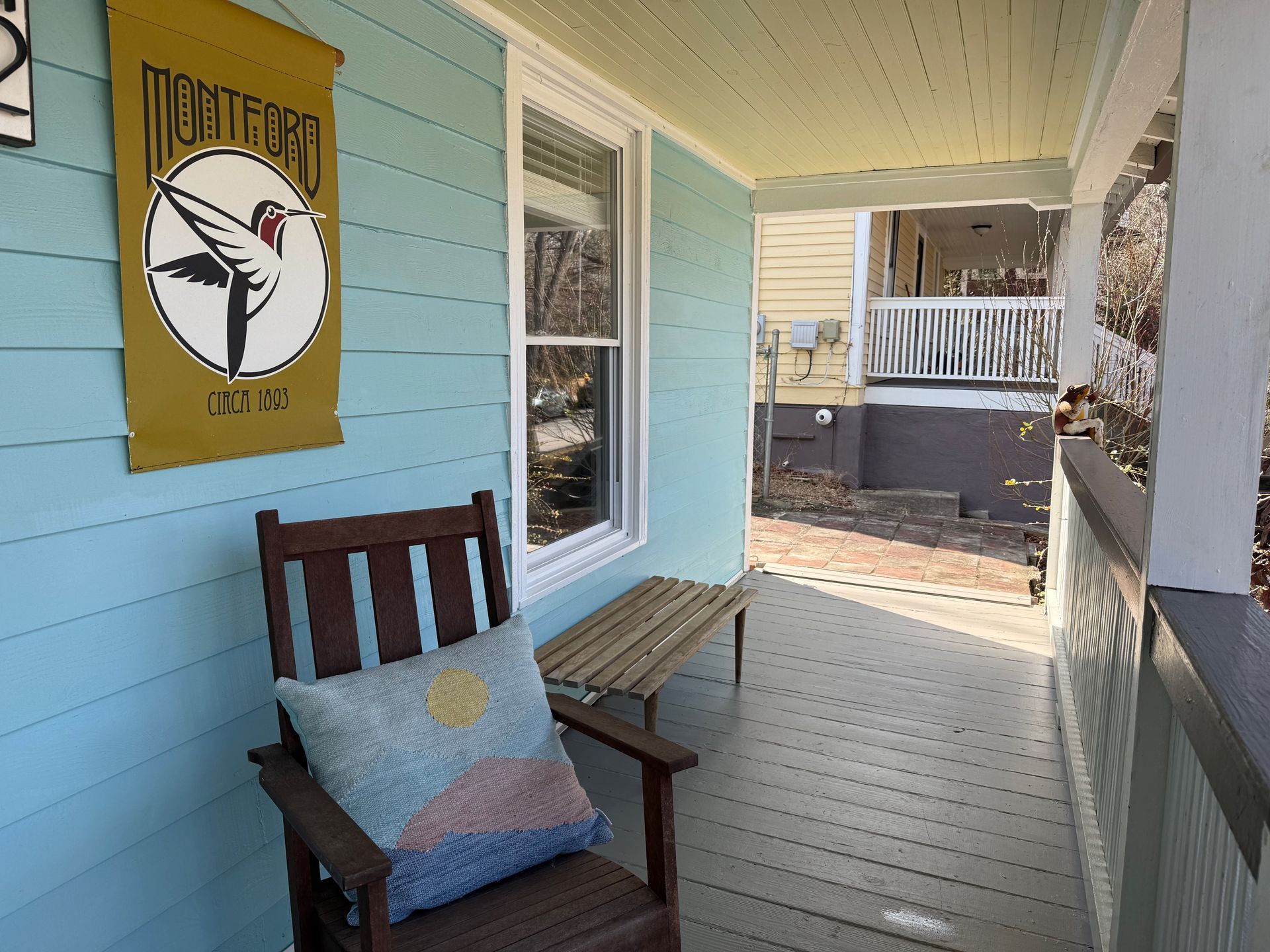 Covered porch with aqua siding, wooden chair and bench, and a yellow bird sign on the wall.
