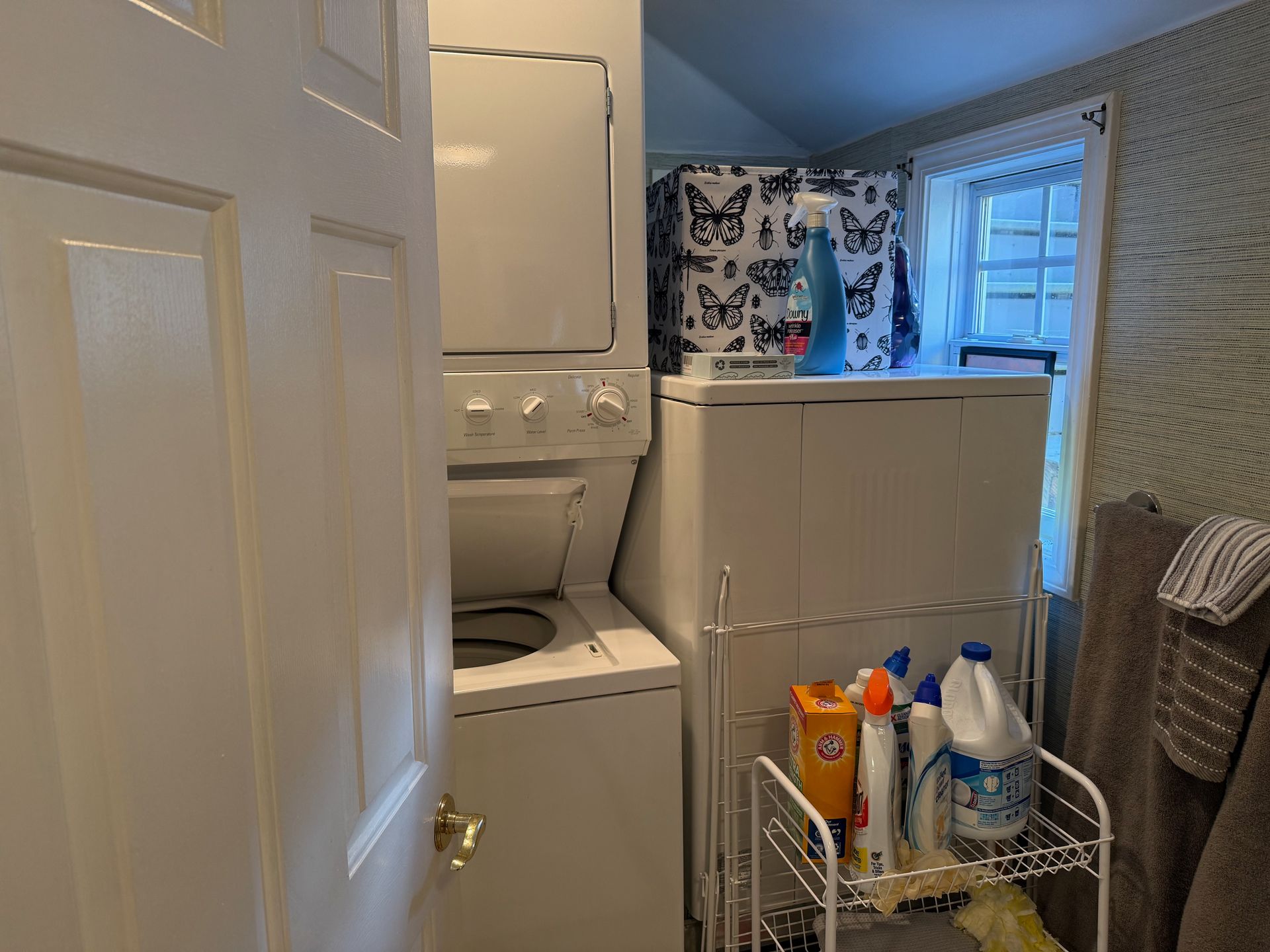Small laundry room with stacked washer-dryer, laundry basket, cleaning supplies, and a window.