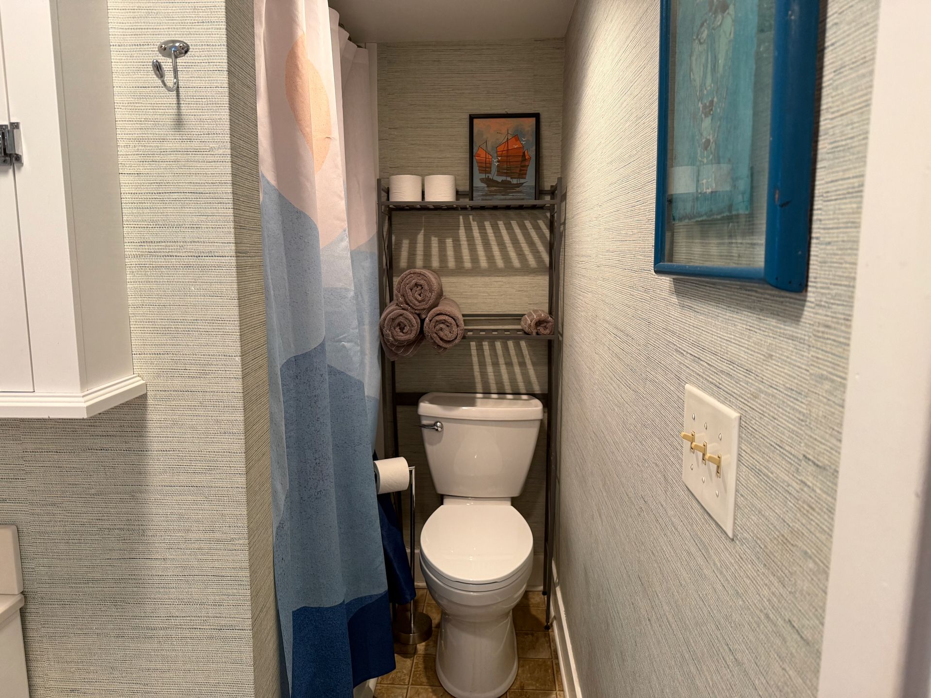 Narrow bathroom with white toilet, blue shower curtain, wall shelf, and textured white walls