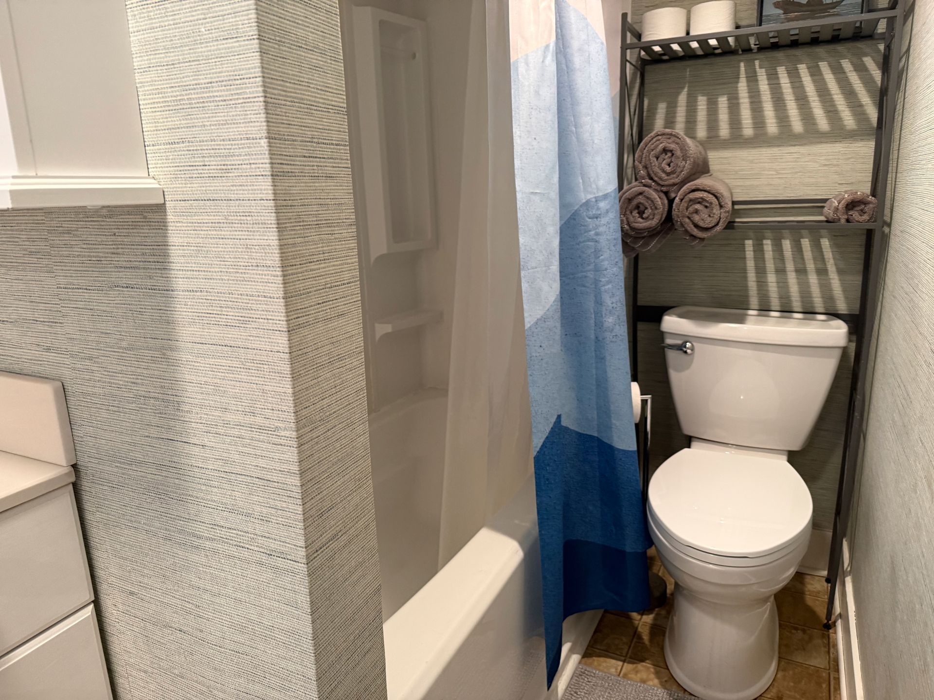 Small bathroom with toilet, black shelving, blue shower curtain, and towel rolls next to a walk-in shower