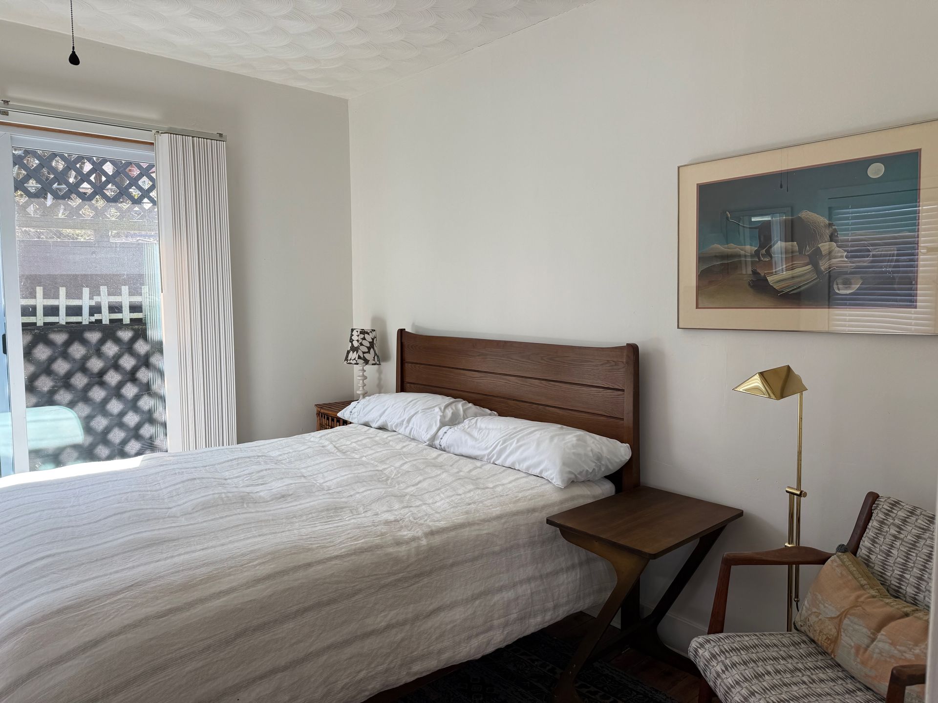 Bright bedroom with white bed, wooden headboard, lamp, chair, and framed wall art by a window.