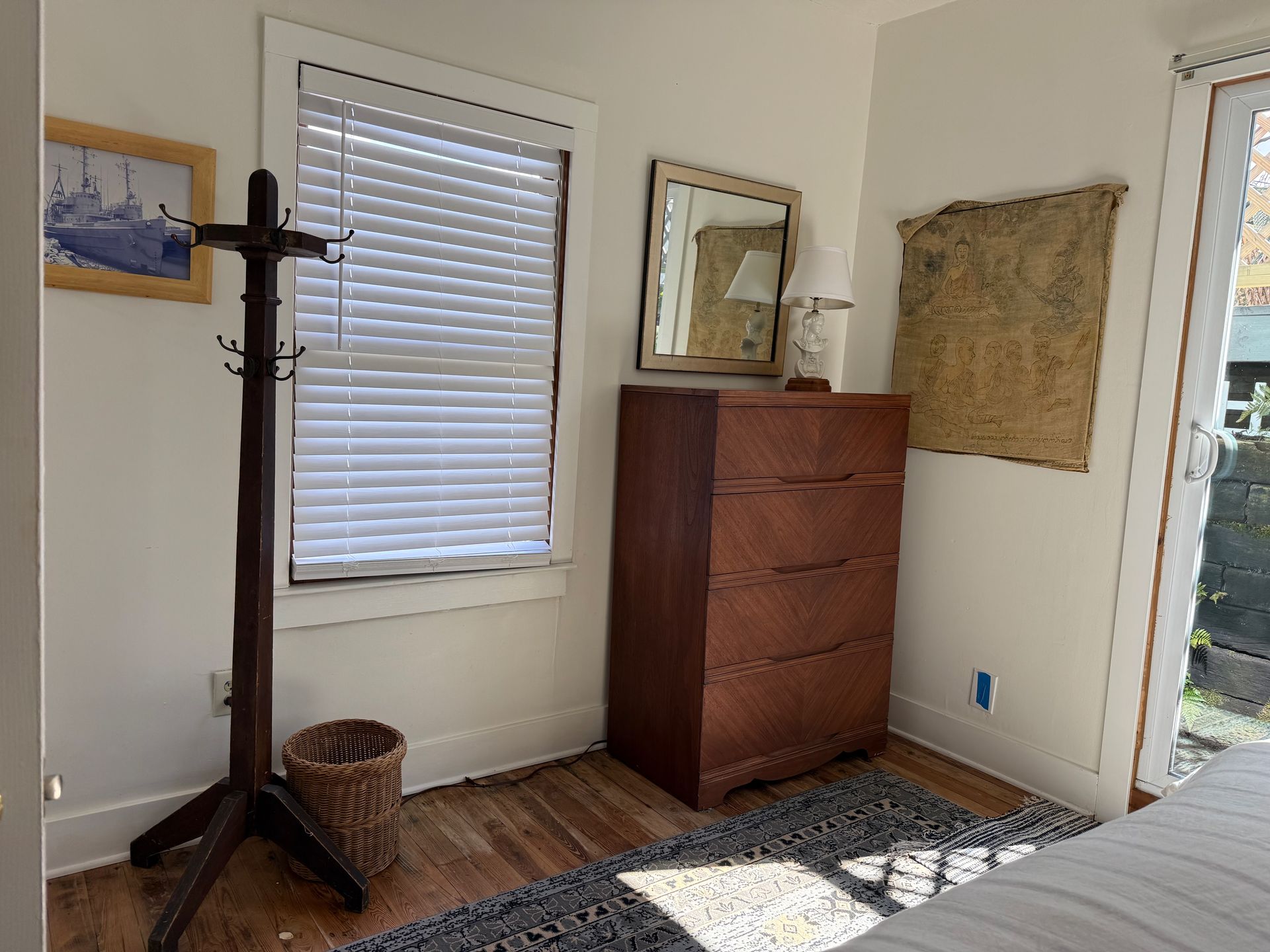 Sparse bedroom with wooden dresser, coat rack, window blinds, woven basket, and rug on hardwood floor