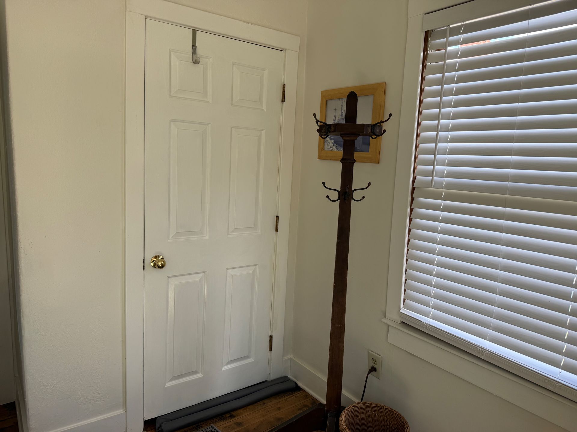 Small room corner with white door, window blinds, and wooden coat rack with hooks.