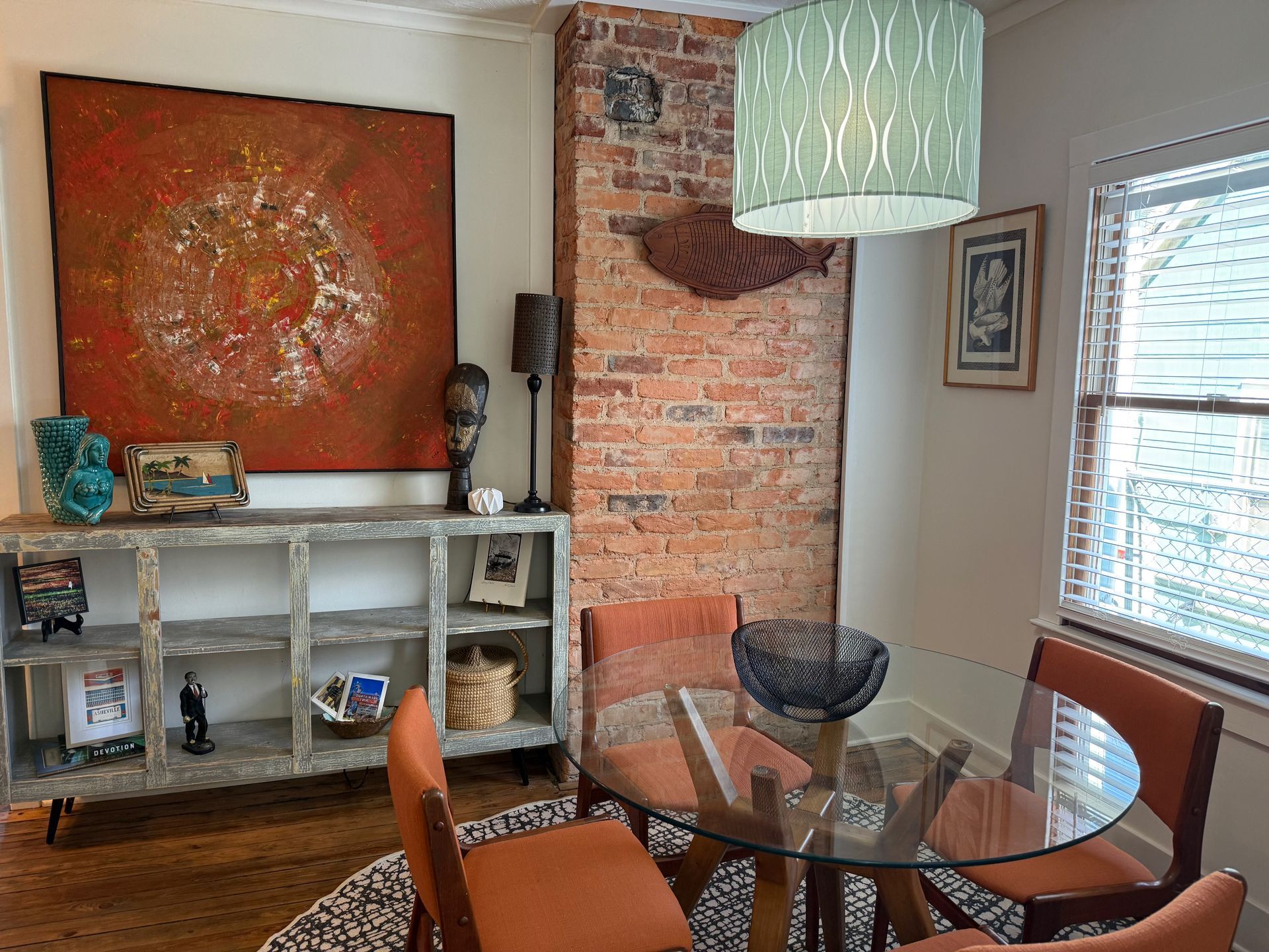 Modern dining nook with glass table, woven chairs, brick wall, white console, and orange artwork