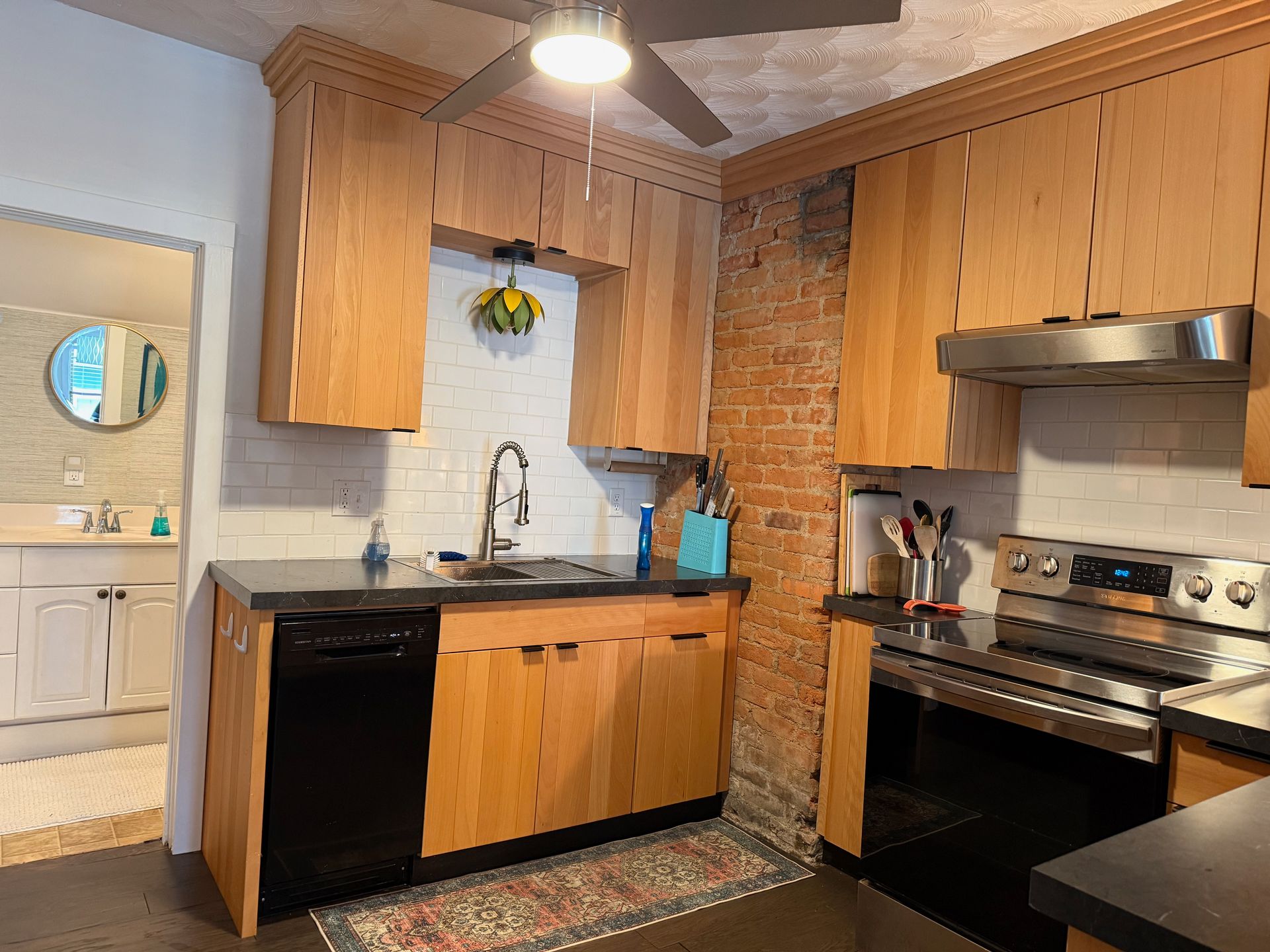 Compact kitchen with wood cabinets, black appliances, brick accent wall, and round mirror by the doorway
