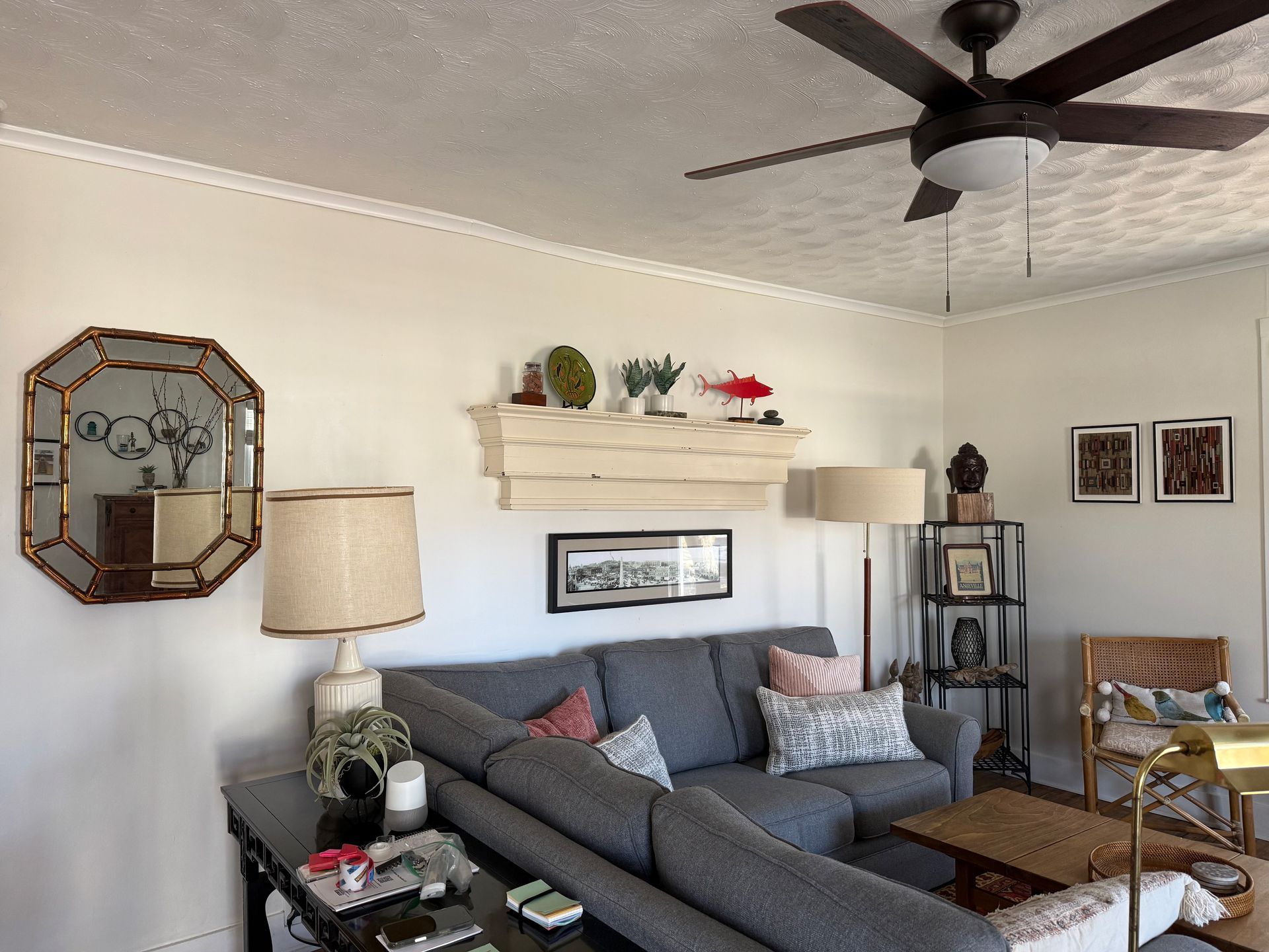 Cozy living room with gray sectional, ceiling fan, lamps, wall art, and shelving on cream walls