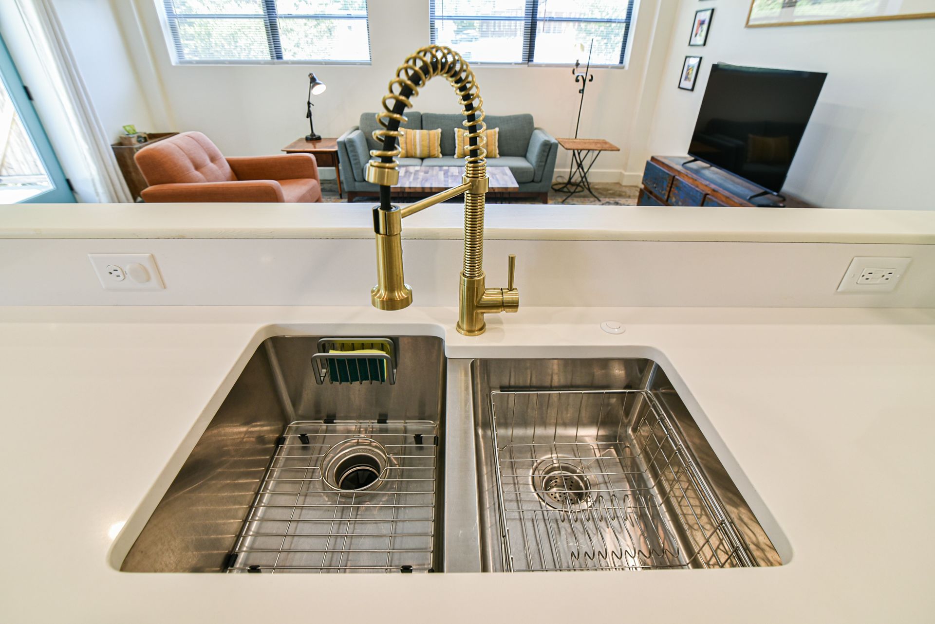 Modern kitchen sink with brass faucet, deep basins, and a living room visible beyond the counter.