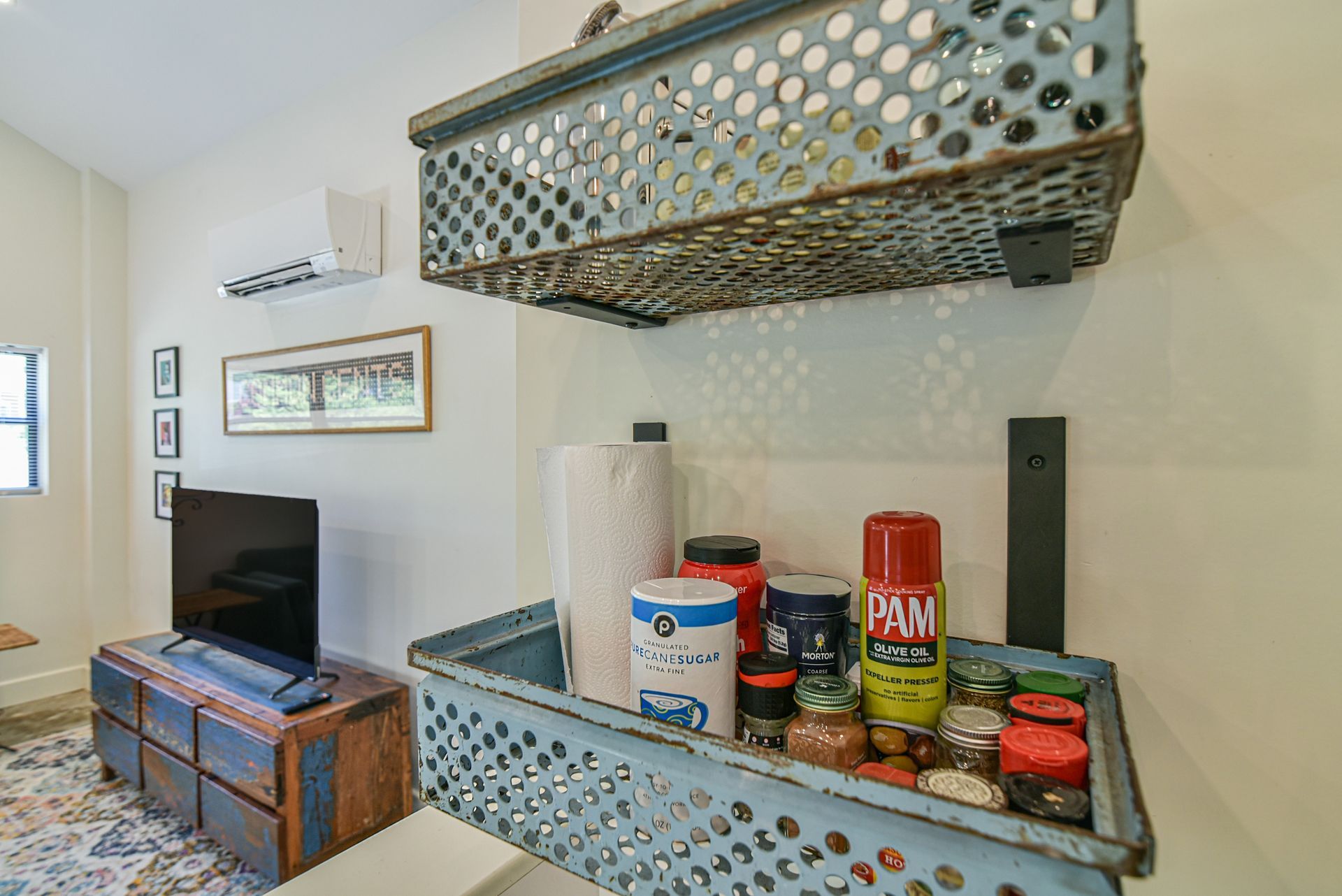 Living room with wall-mounted metal shelves holding jars and decor above a TV and wooden chest.