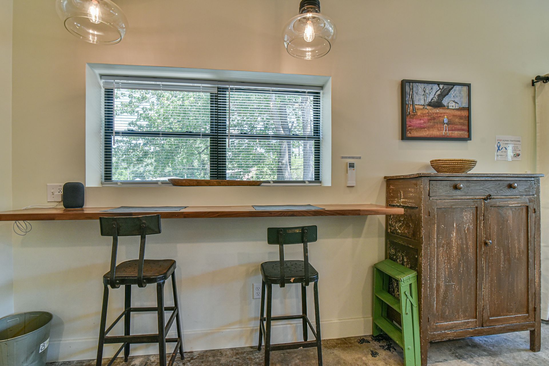 Cafe counter with two stools under a large window and hanging lights, beside a rustic wooden cabinet