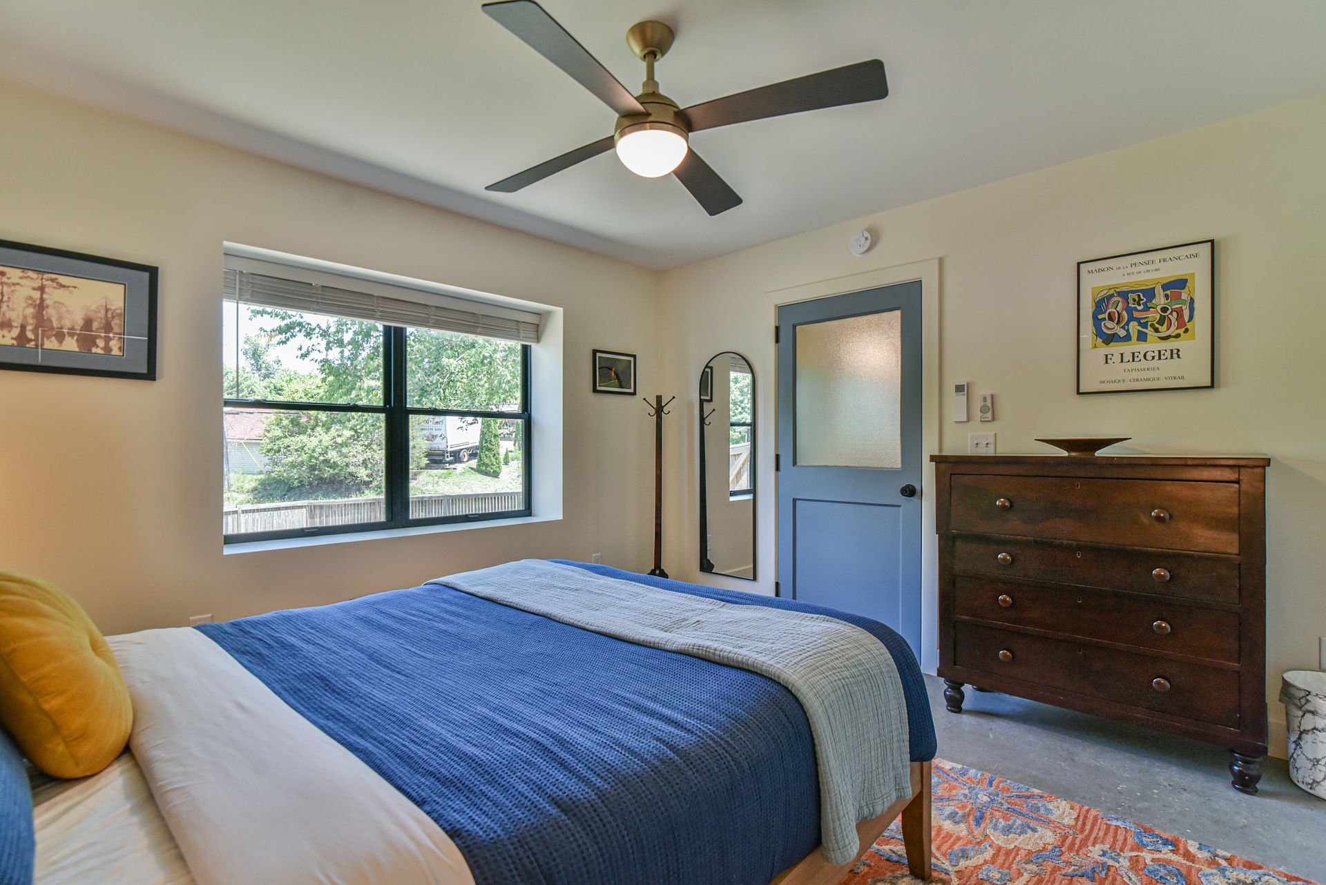 Bright bedroom with blue bedspread, window, ceiling fan, dresser, and colorful rug