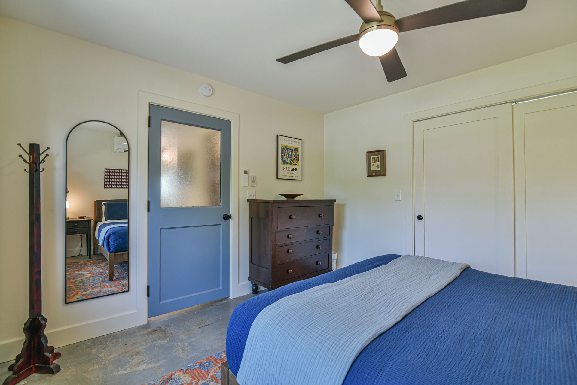 Bedroom with blue bedspread, blue door, dresser, mirror, and ceiling fan.