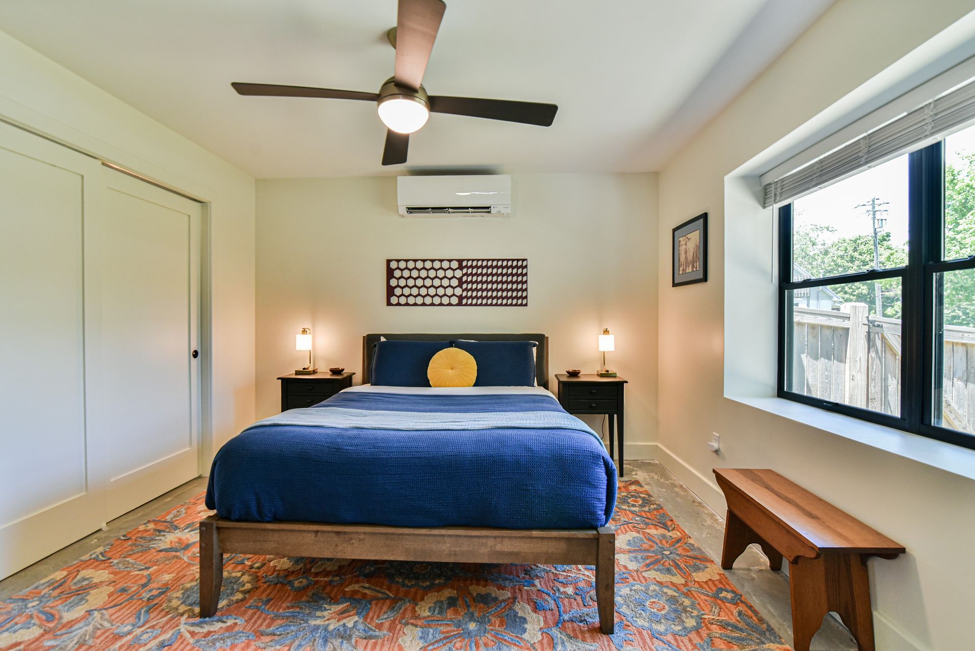 Bedroom with blue bedspread, patterned rug, ceiling fan, nightstands, and wall art.