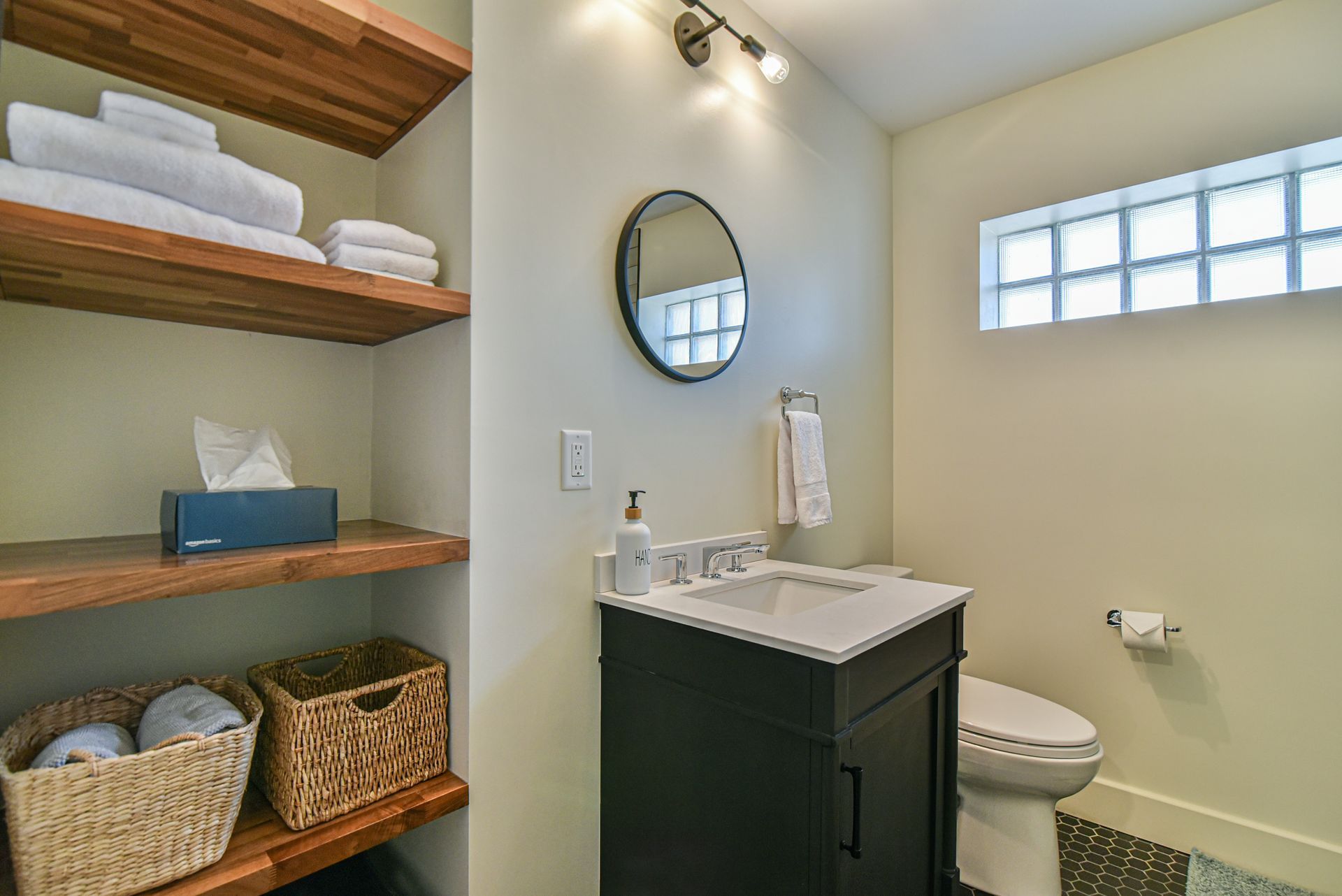 Small bathroom with dark vanity, round mirror, shelves, toilet, and woven baskets￼