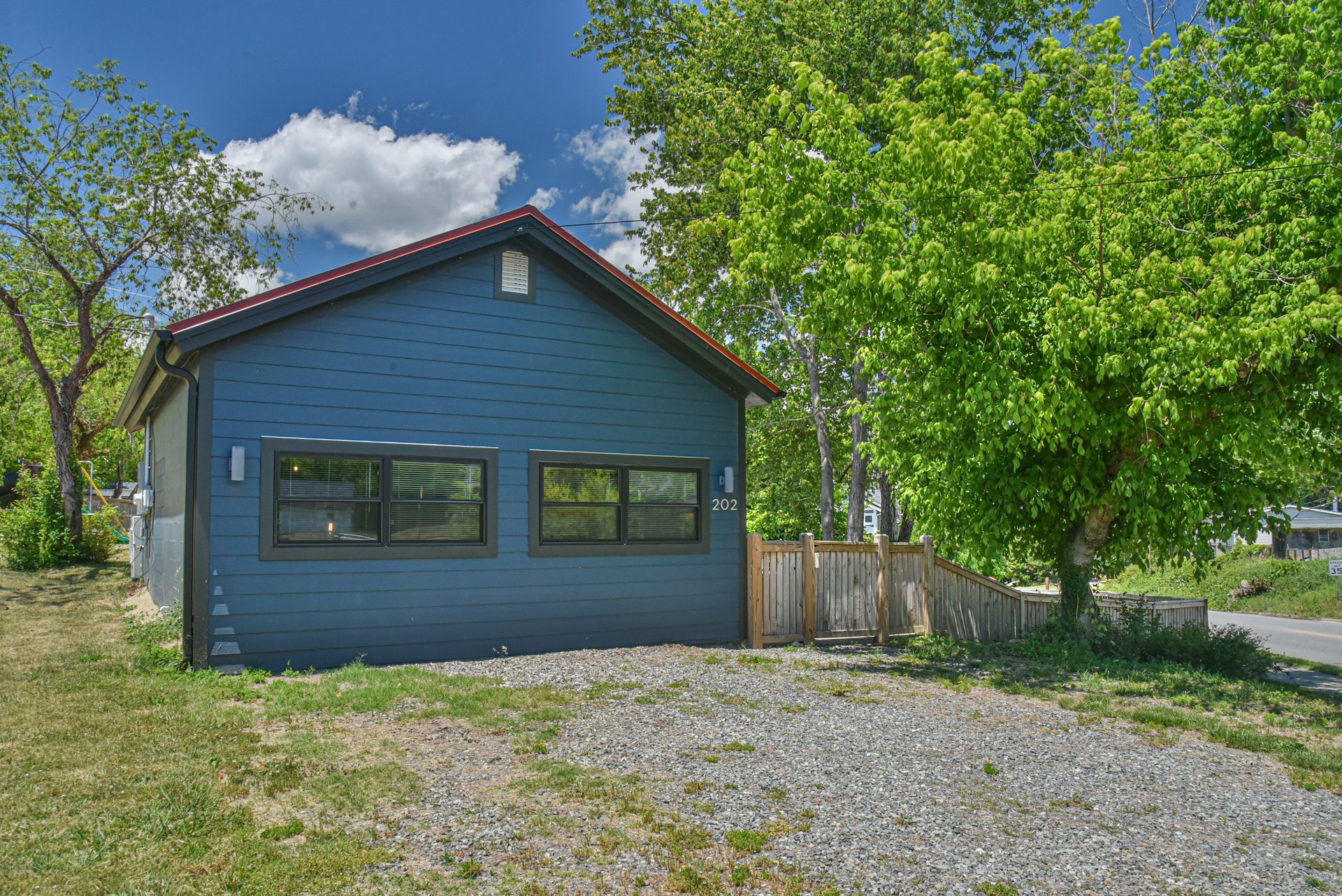 Blue house with green trim and gravel driveway under bright sky, surrounded by trees