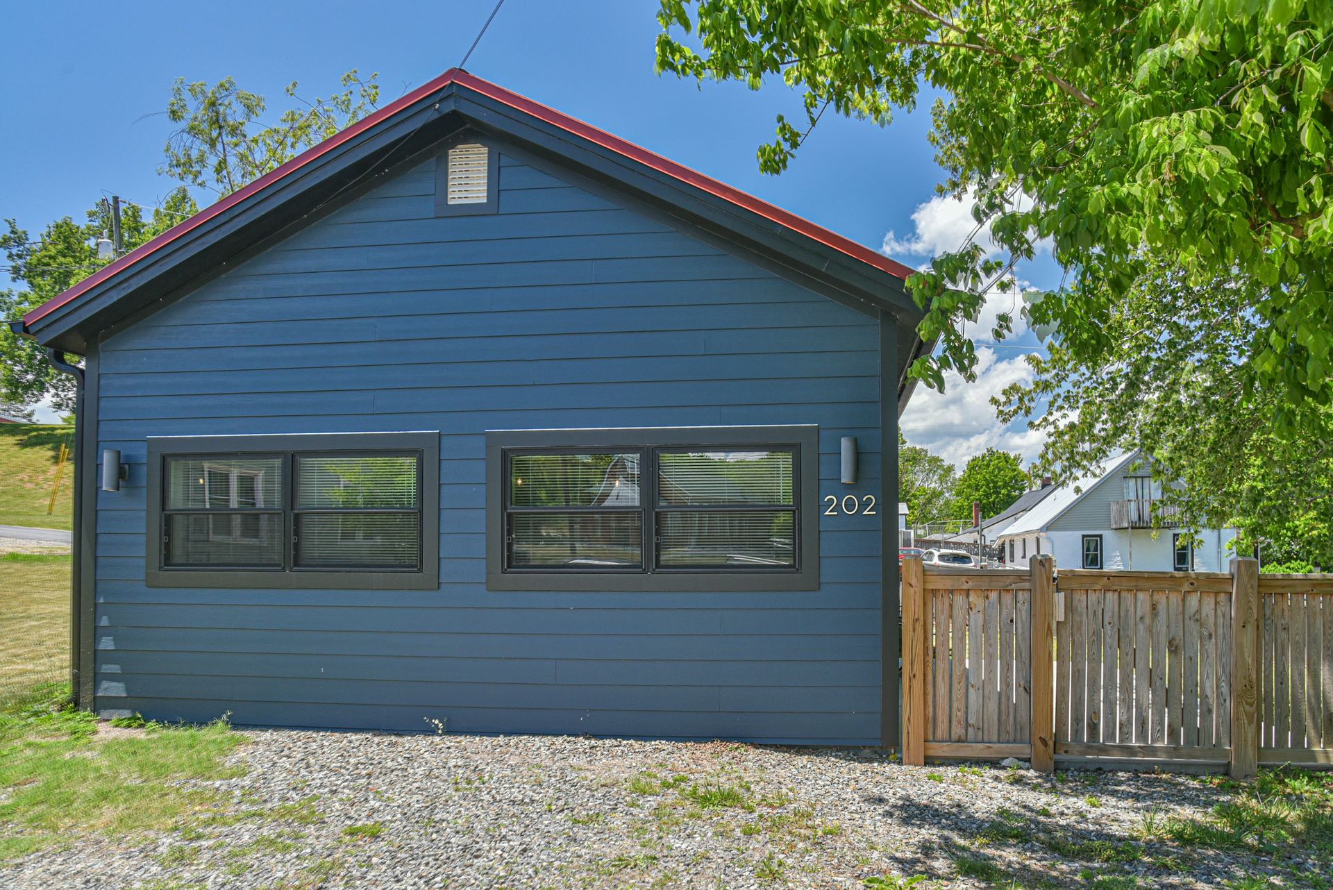 Blue single-story house with two front windows, gravel yard, and a wooden fence under a sunny sky
