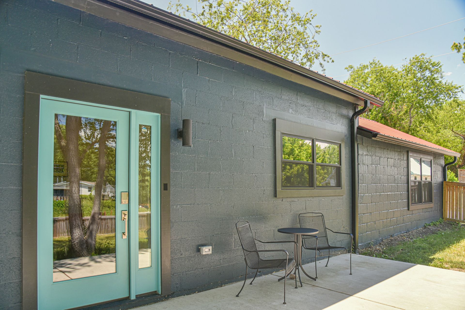 Gray house exterior with turquoise door, black window, and small patio table and chairs under trees
