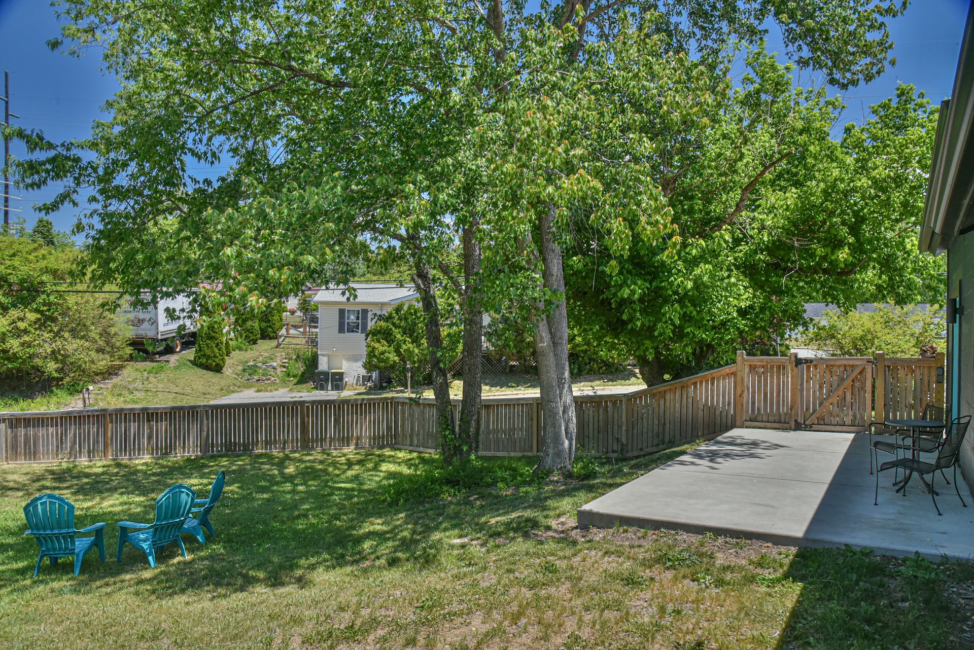 Backyard with green lawn, large trees, wooden fence, patio, and blue chairs in sunlight